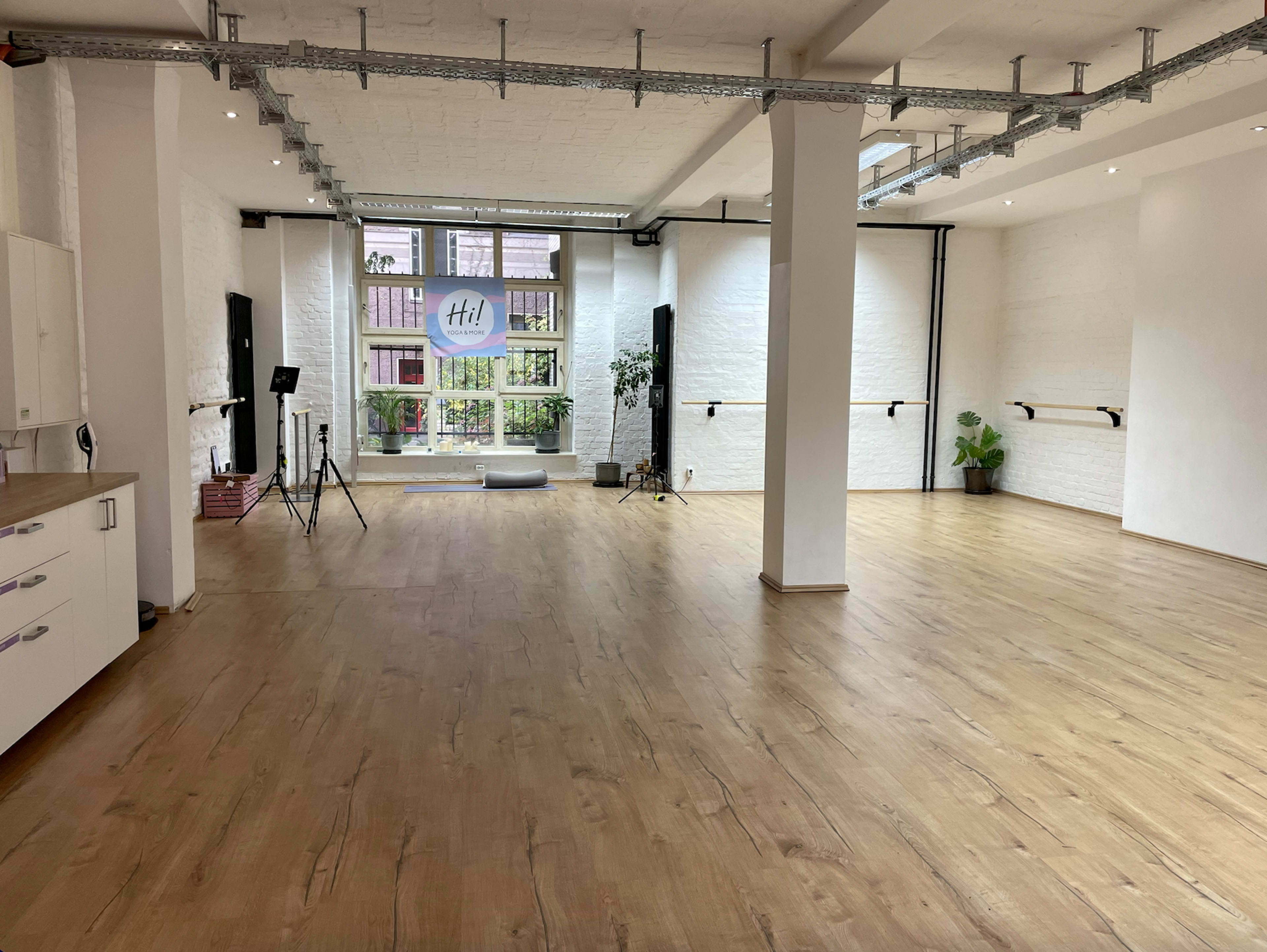 A spacious, well-lit dance studio featuring wooden flooring and large windows with a view of greenery outside.