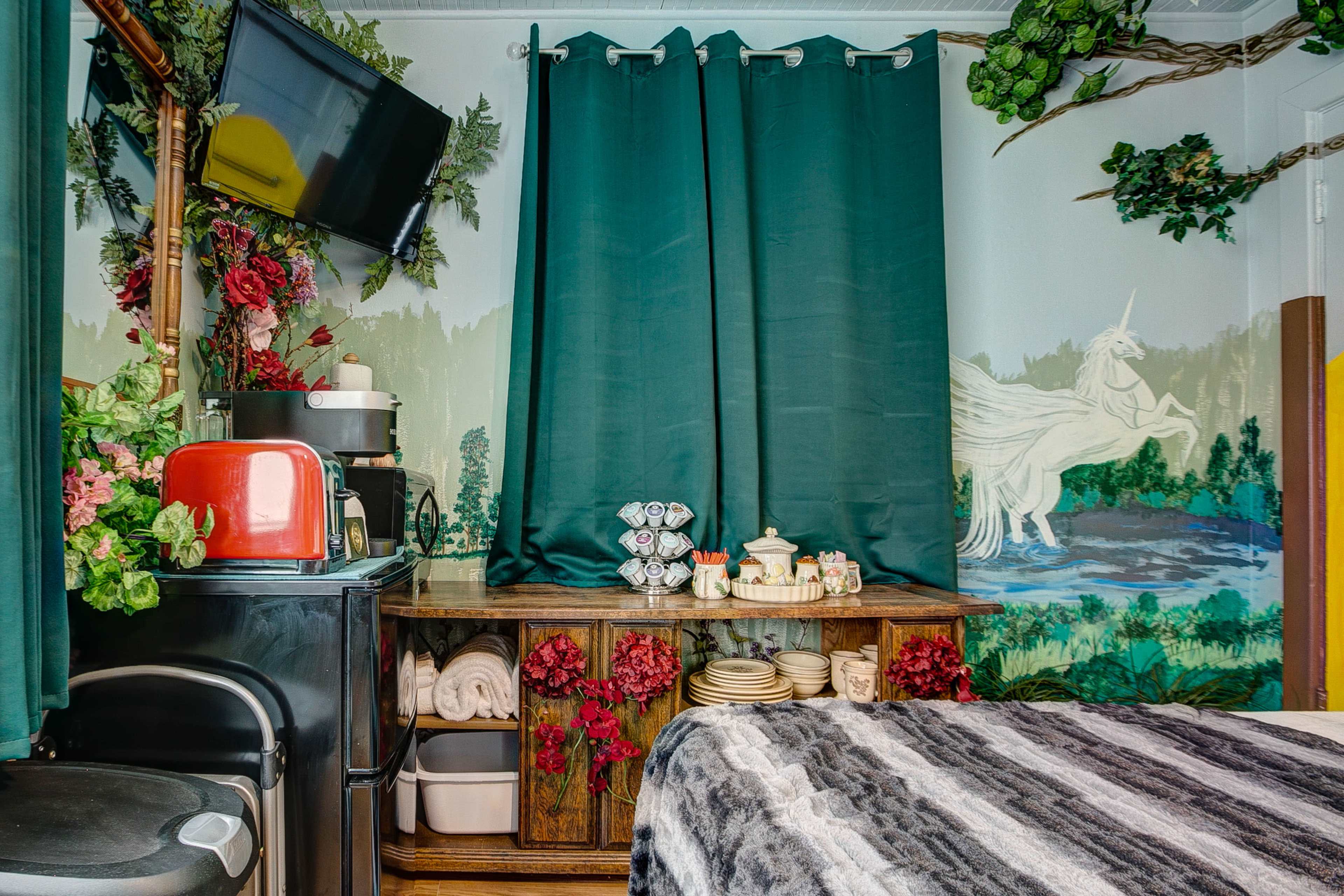 A cozy room with dark green curtains, a TV mounted on the wall, a wooden shelf adorned with floral decorations, and a mural of a unicorn in a forest setting.