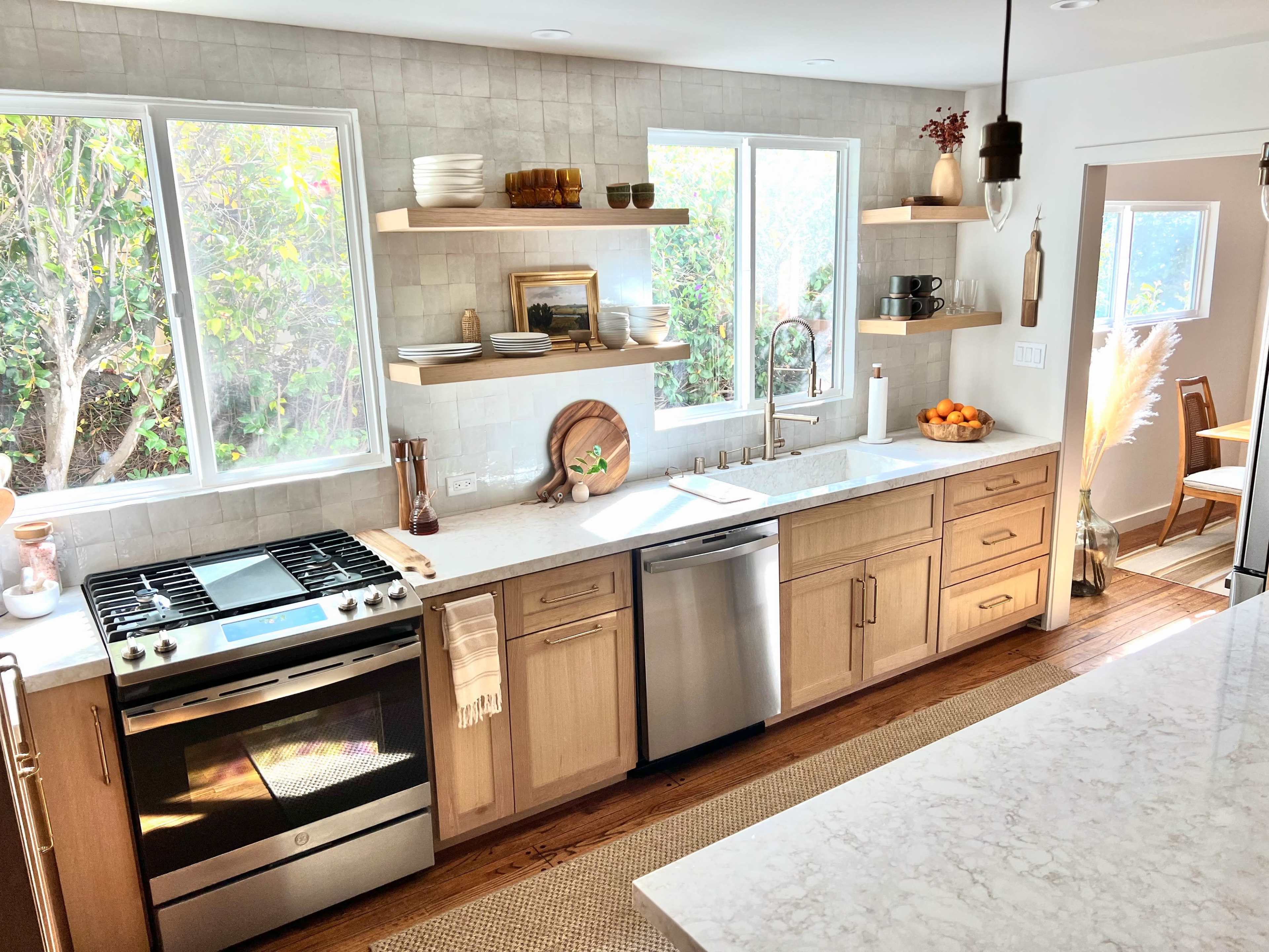 A bright kitchen features wooden shelves with decorative items, a stainless steel stove, and a large marble countertop with a bowl of oranges.