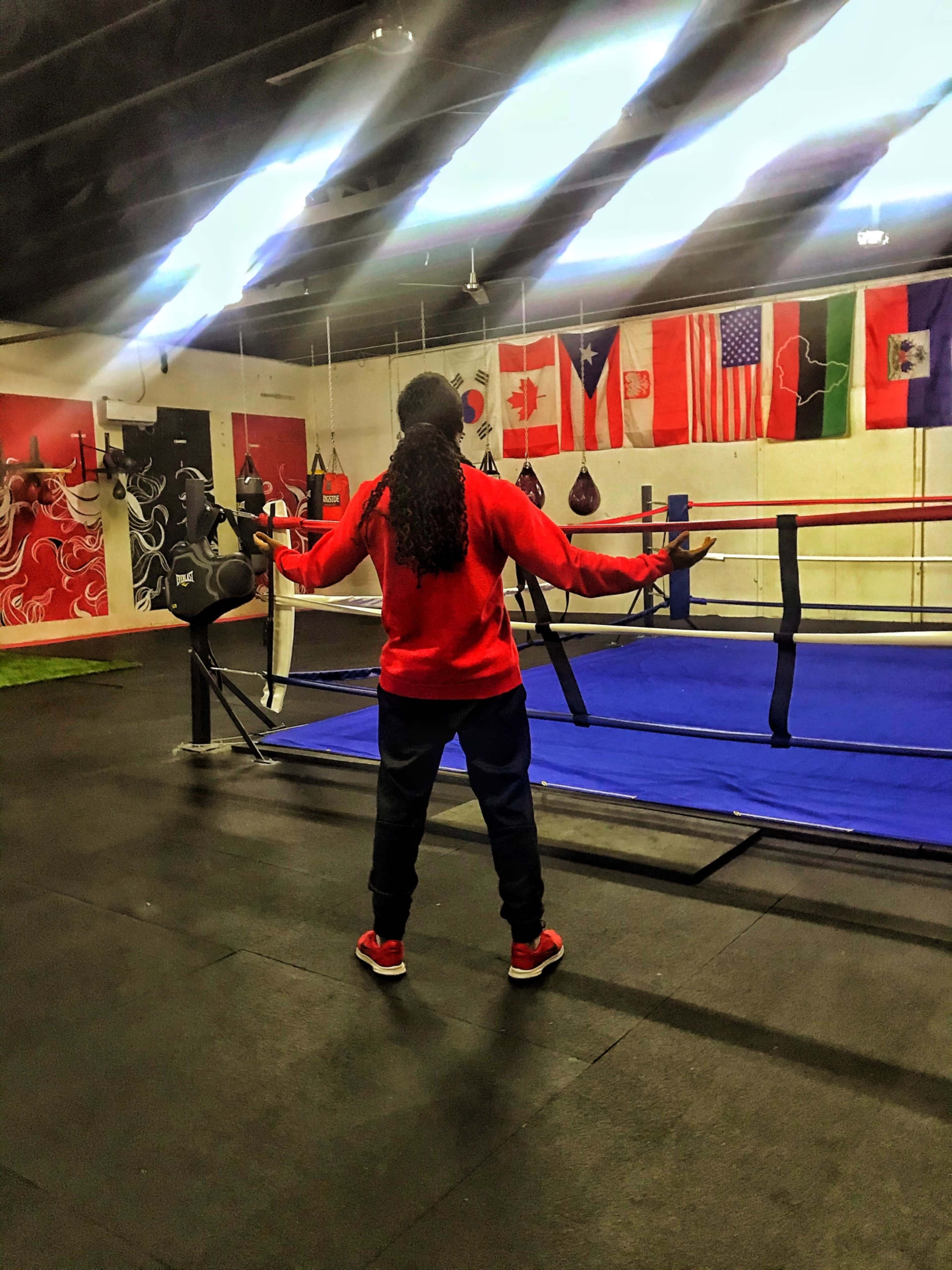 A person in a red sweatshirt and dark pants stands with arms outstretched in a boxing gym, facing a blue ring surrounded by flags on the walls.