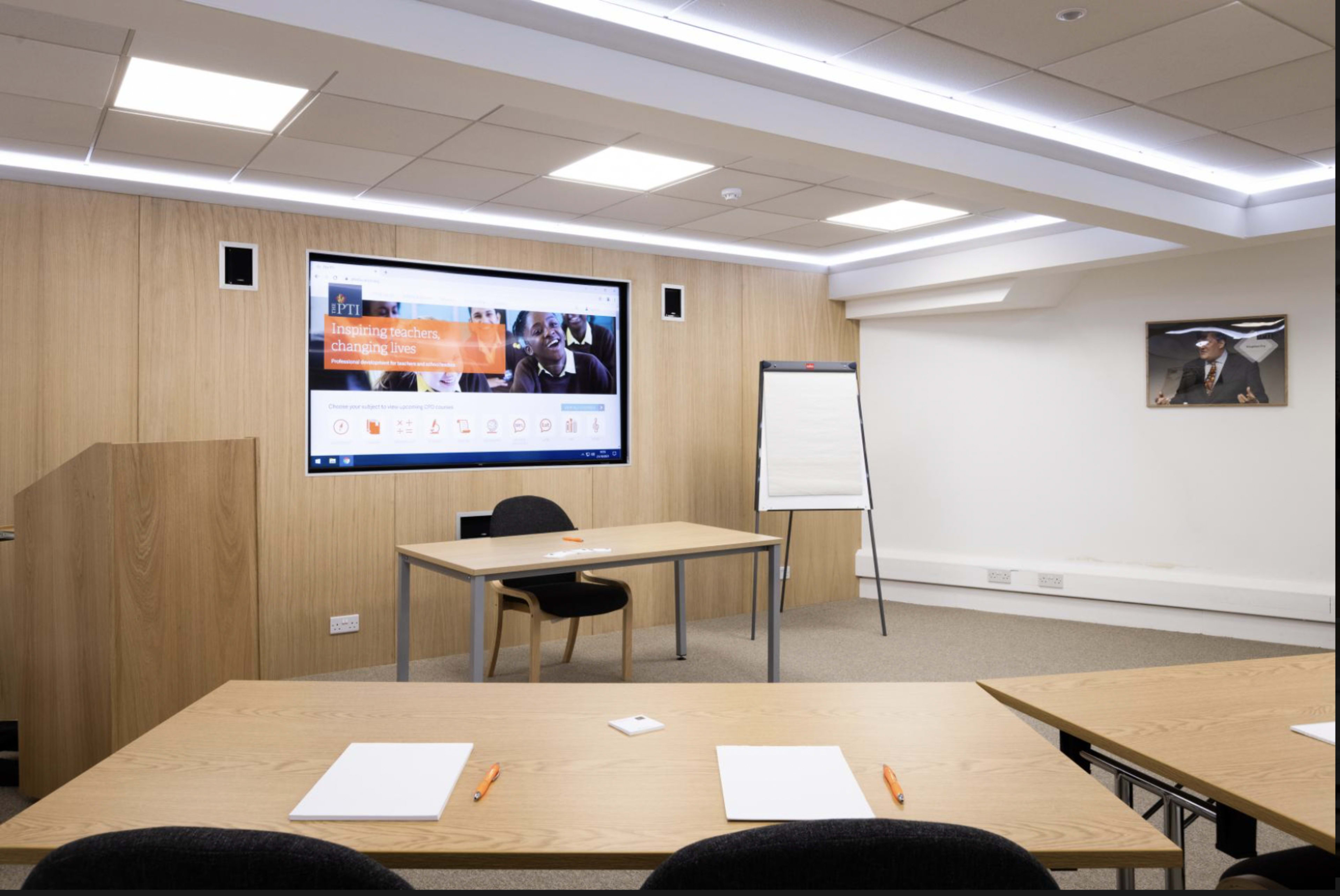 A modern meeting room featuring a large screen, a wooden presentation podium, a table with chairs, and a whiteboard.