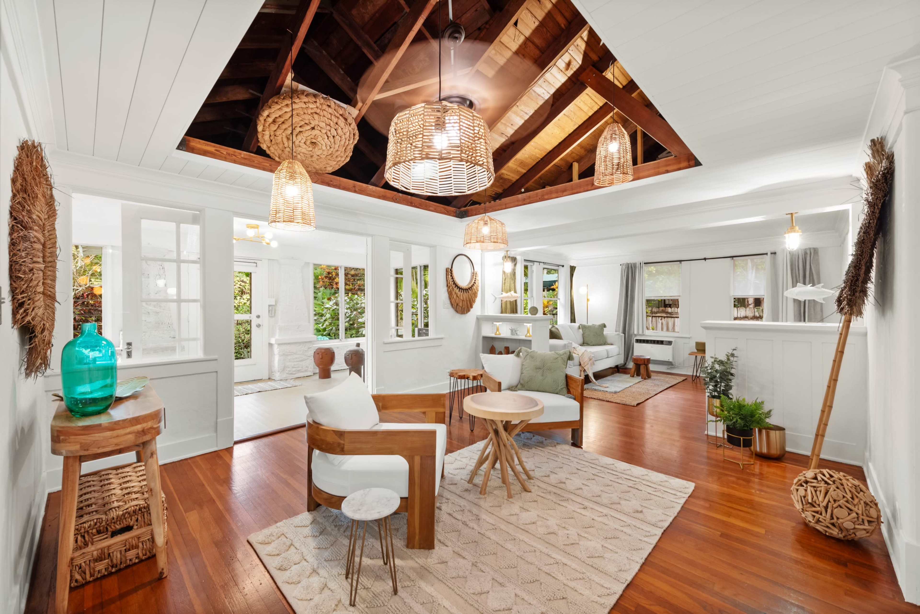 The image shows a bright, open living space with wooden floors, a high ceiling featuring exposed beams, and a mix of decorative furniture and plants.