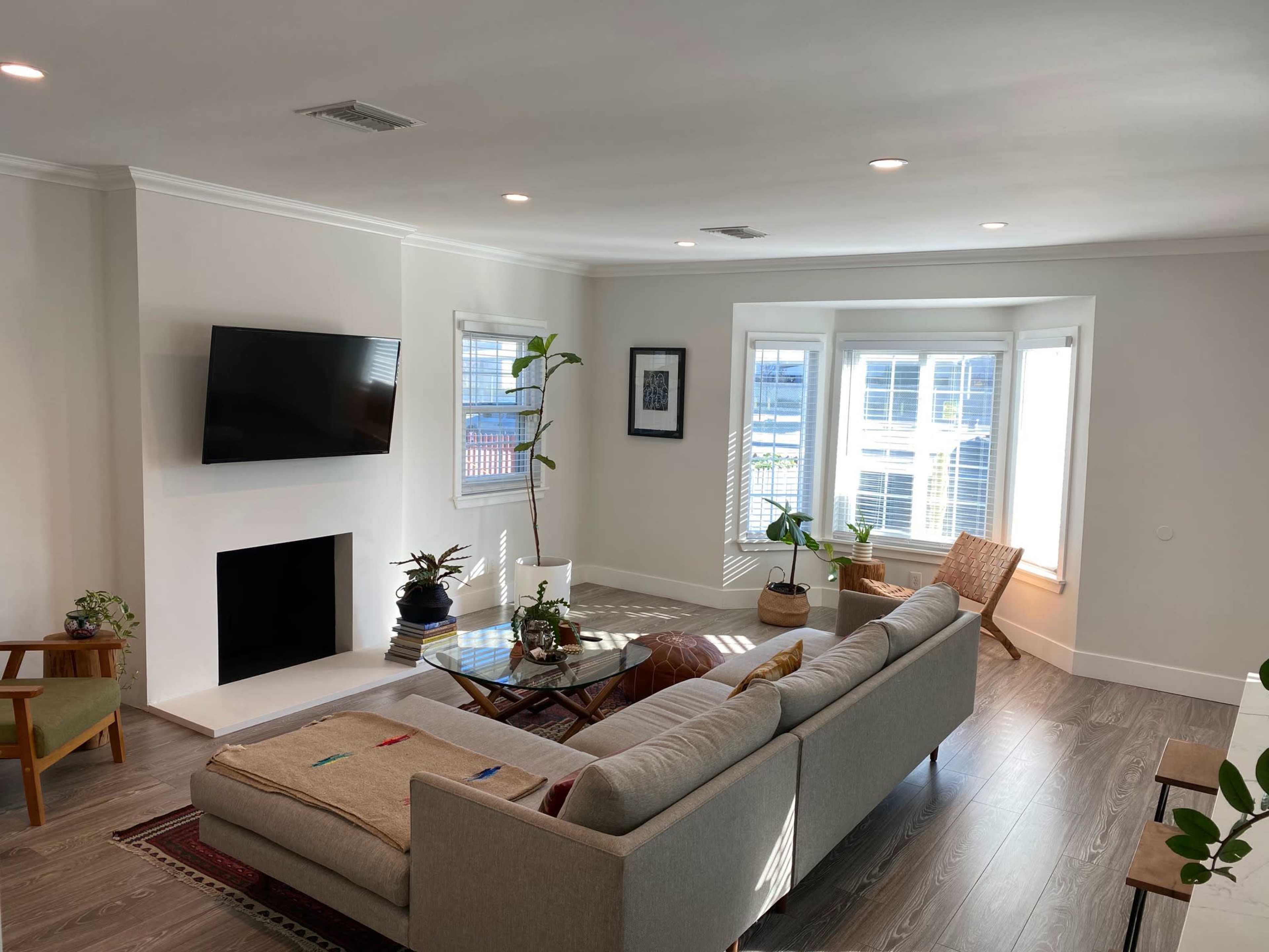 A modern living room features a large gray sectional sofa, a flat-screen TV mounted on the wall, and large windows allowing natural light to fill the space.