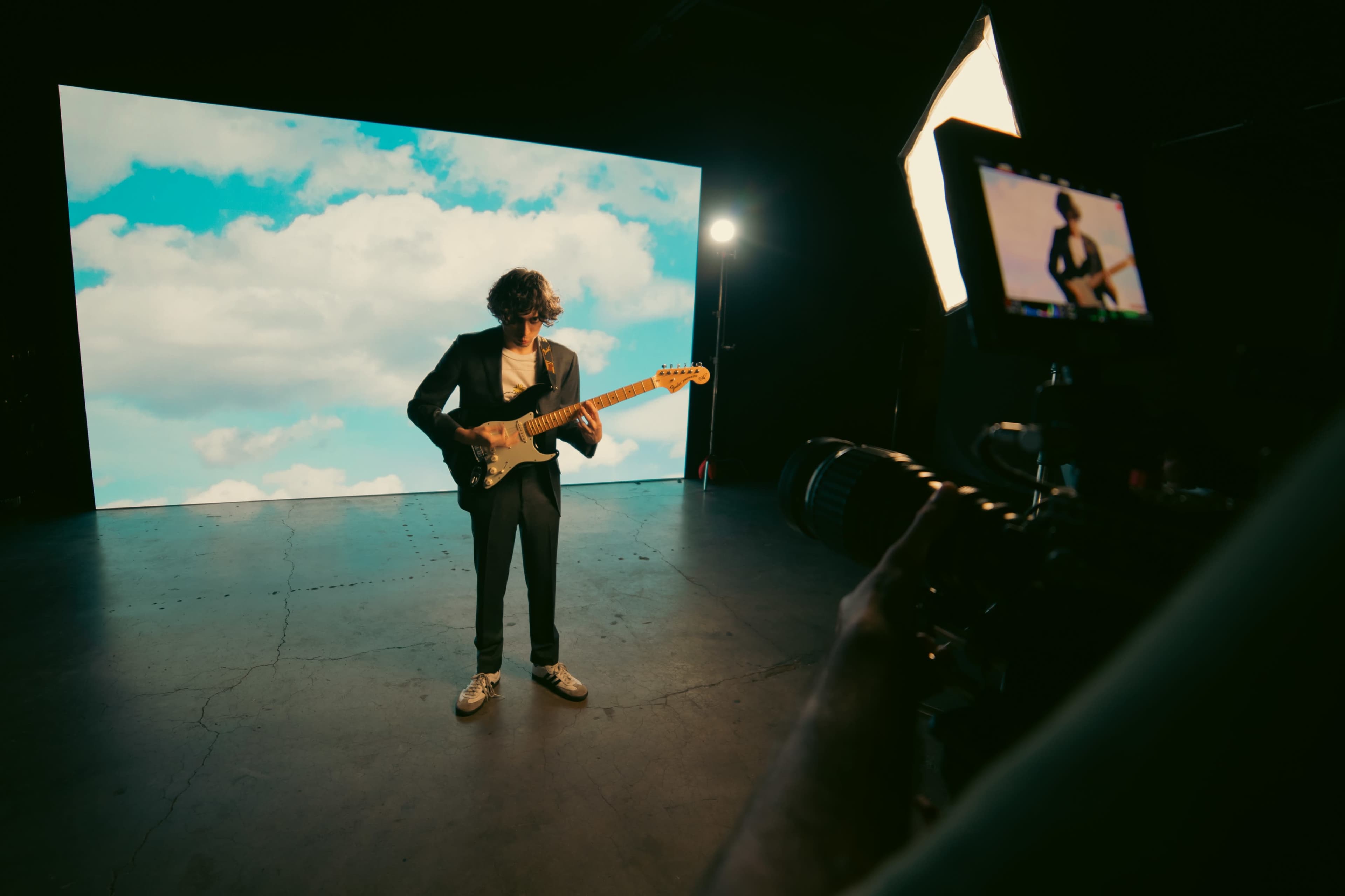 A musician stands on stage with an electric guitar in front of a large screen displaying a cloudy sky.