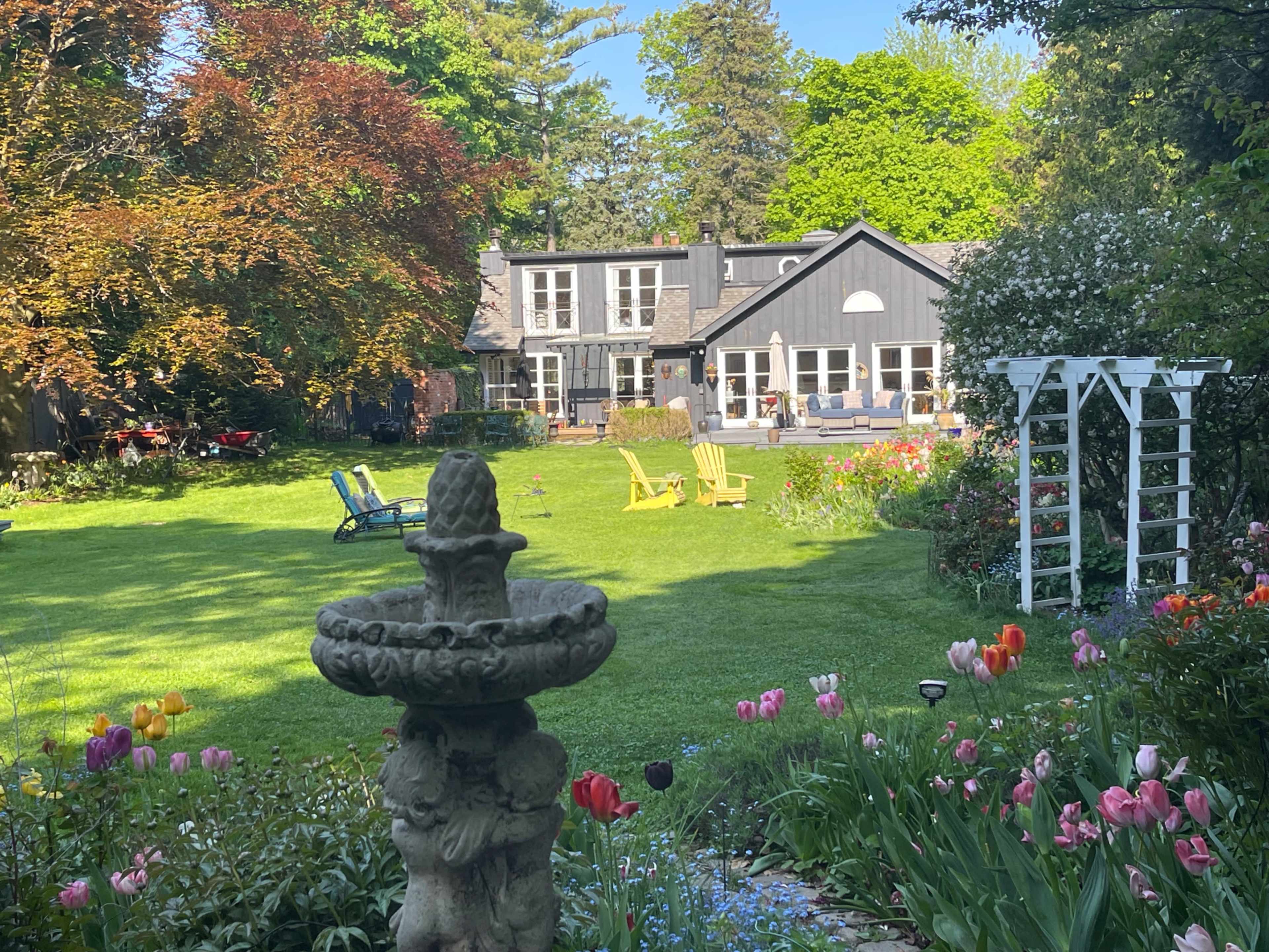 A well-maintained garden features colorful flowers and a stone fountain, with a house and seating area visible in the background.
