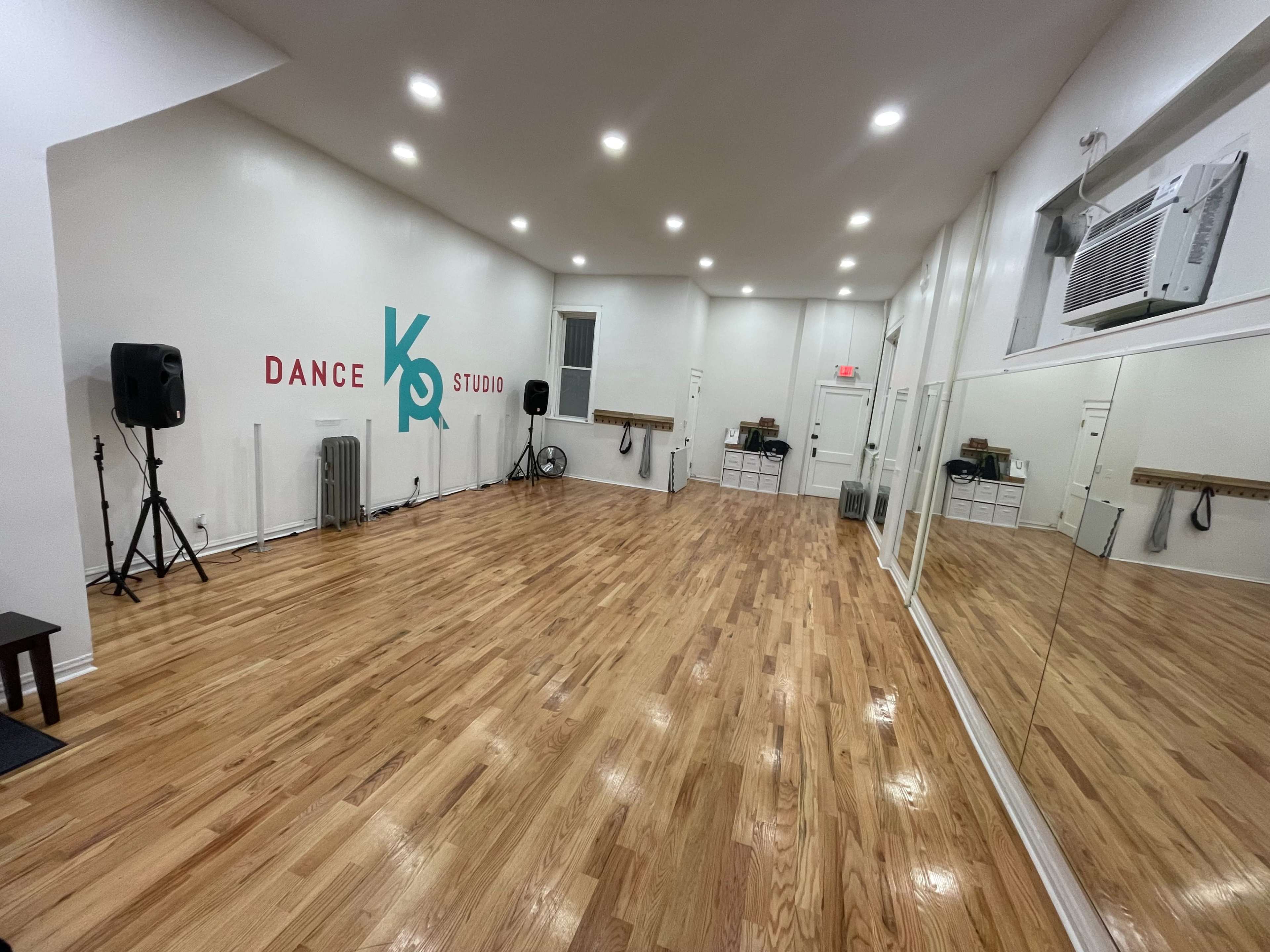 The image shows a spacious dance studio with polished wooden floors, mirrored walls, and a speaker system in one corner.