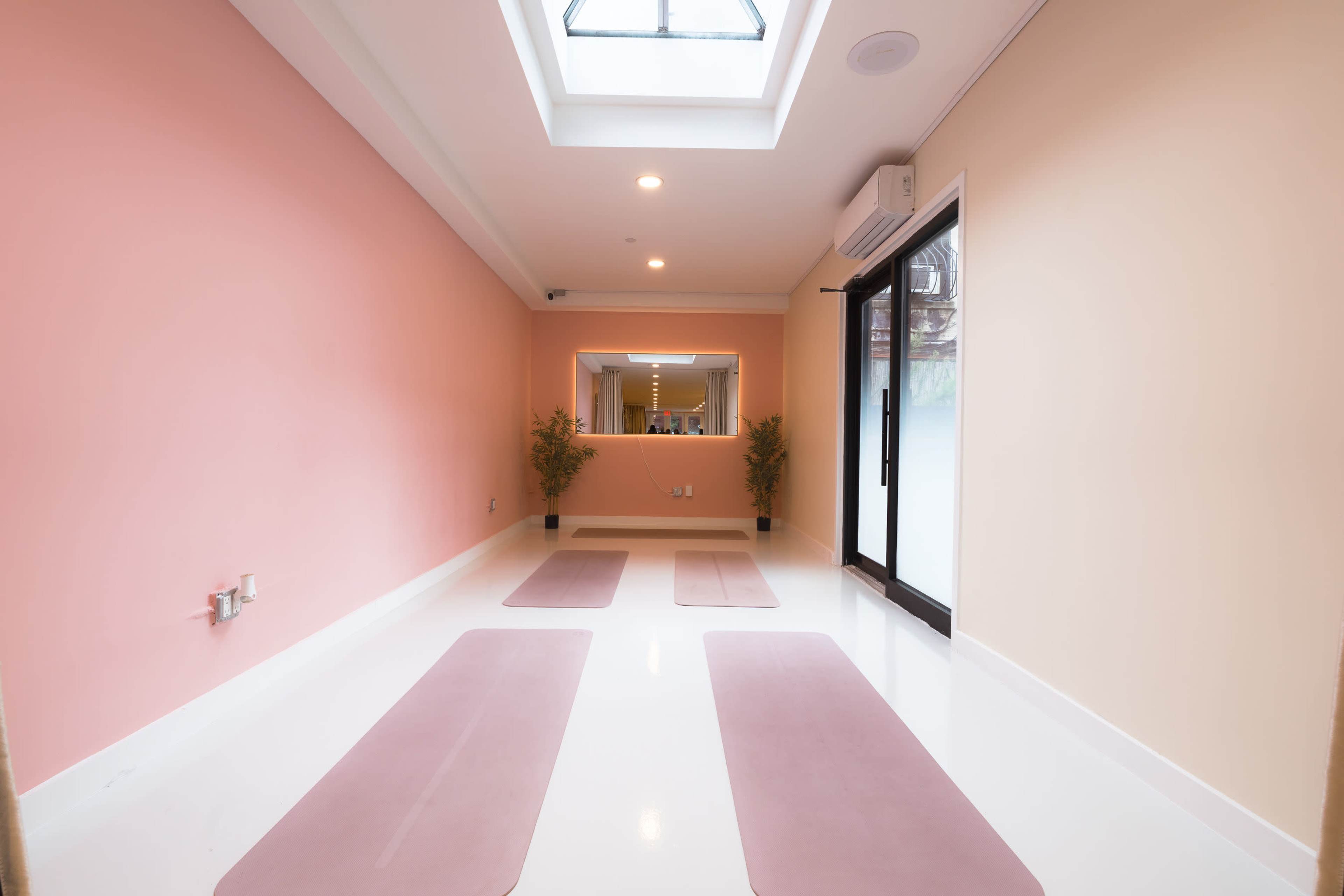 The image shows a tranquil yoga studio with three exercise mats placed on a white floor beneath a skylight and walls painted in soft pink and beige.