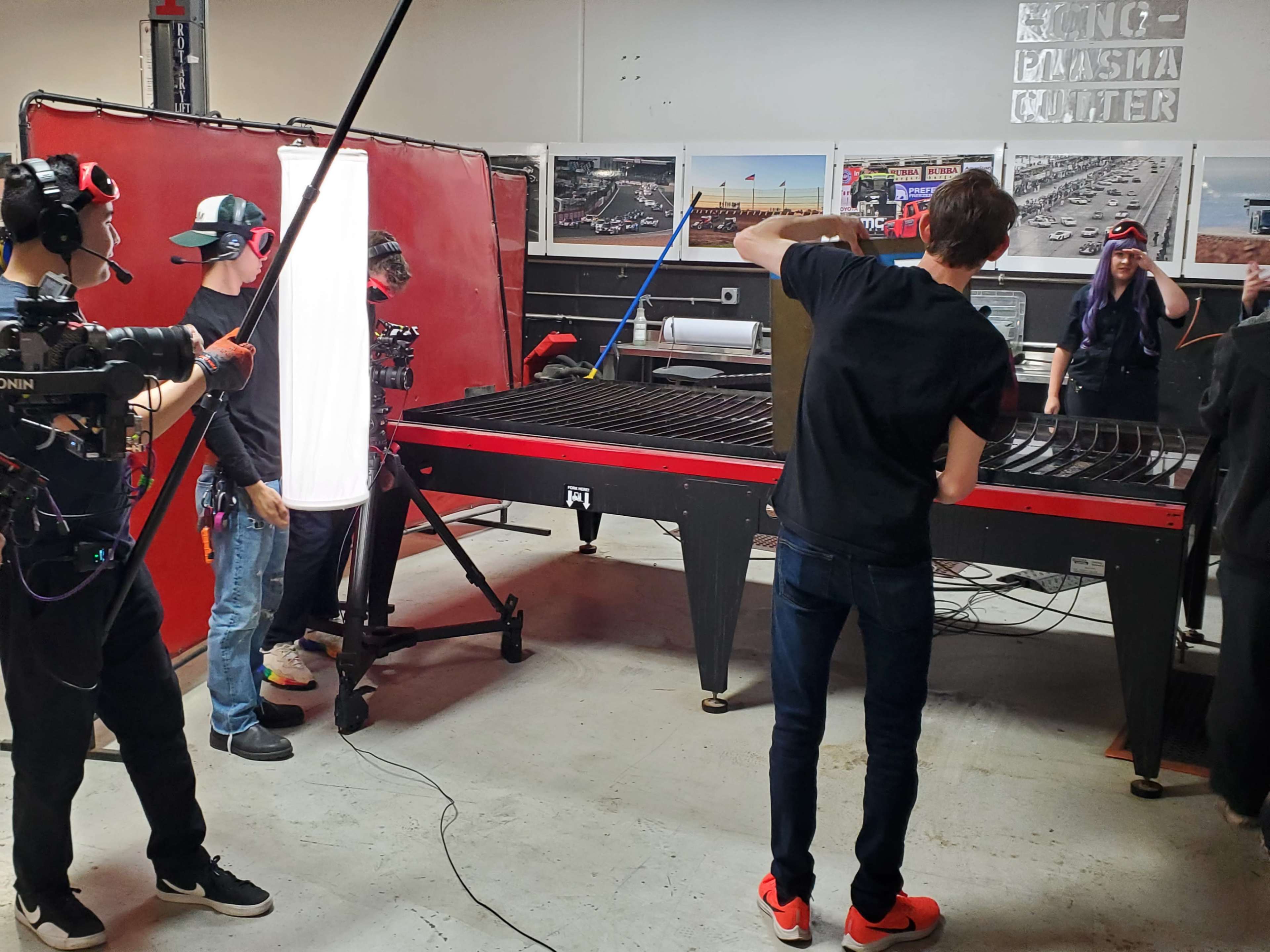 A group of people is filming a demonstration near a large plasma cutter in a workshop setting.