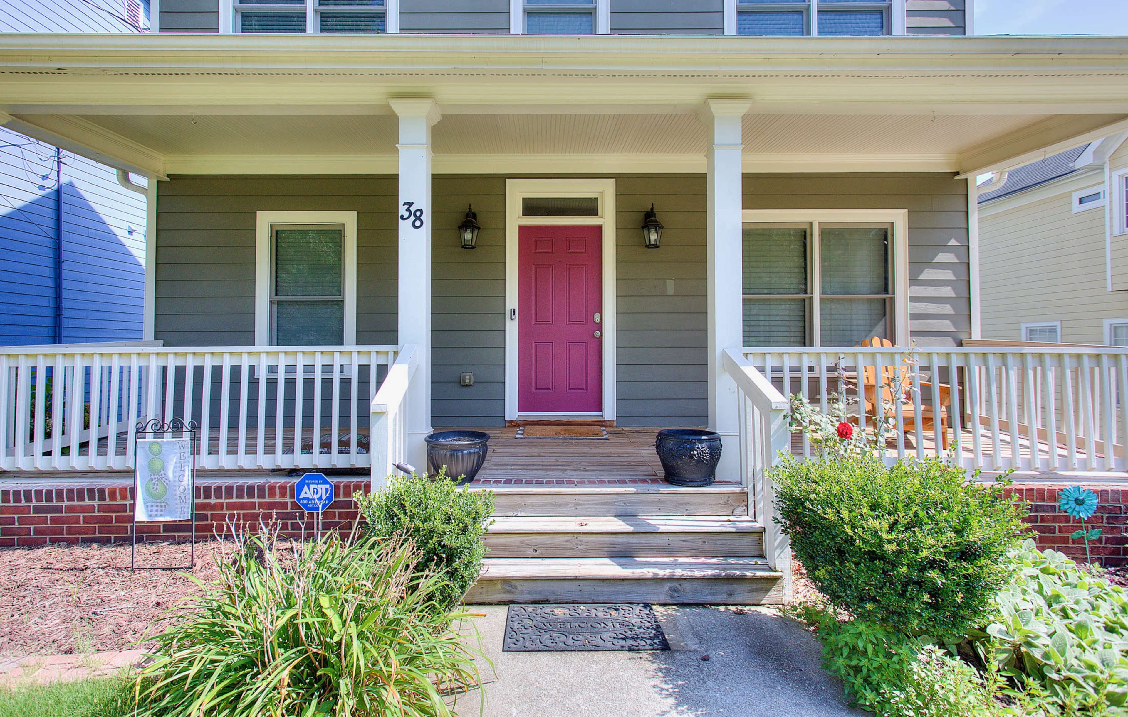 A house entrance with a pink front door, surrounded by a porch with white railings and potted plants on either side.