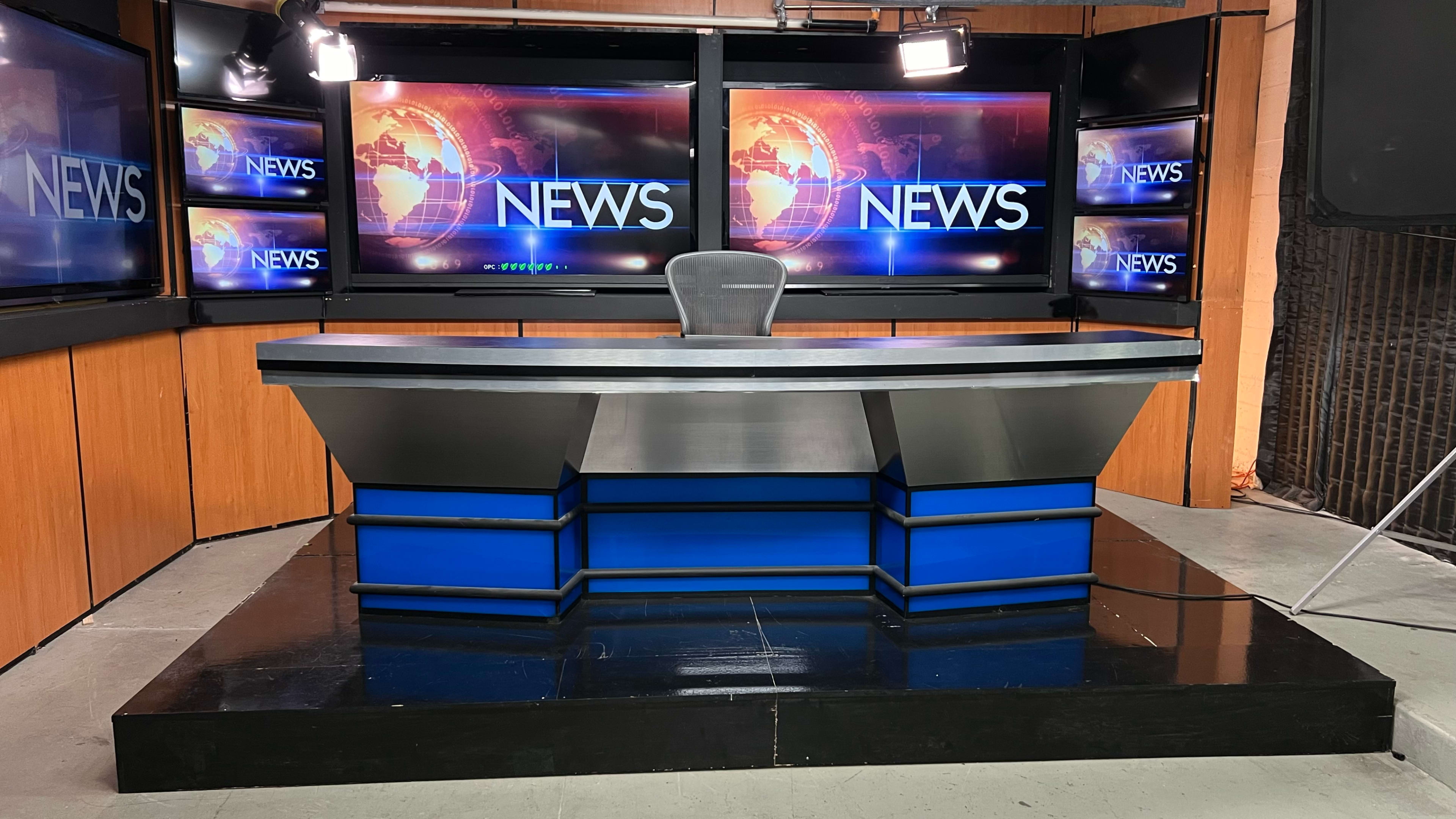 The image shows a news studio set featuring a metallic news desk with a black and blue design, surrounded by screens displaying the word "NEWS."
