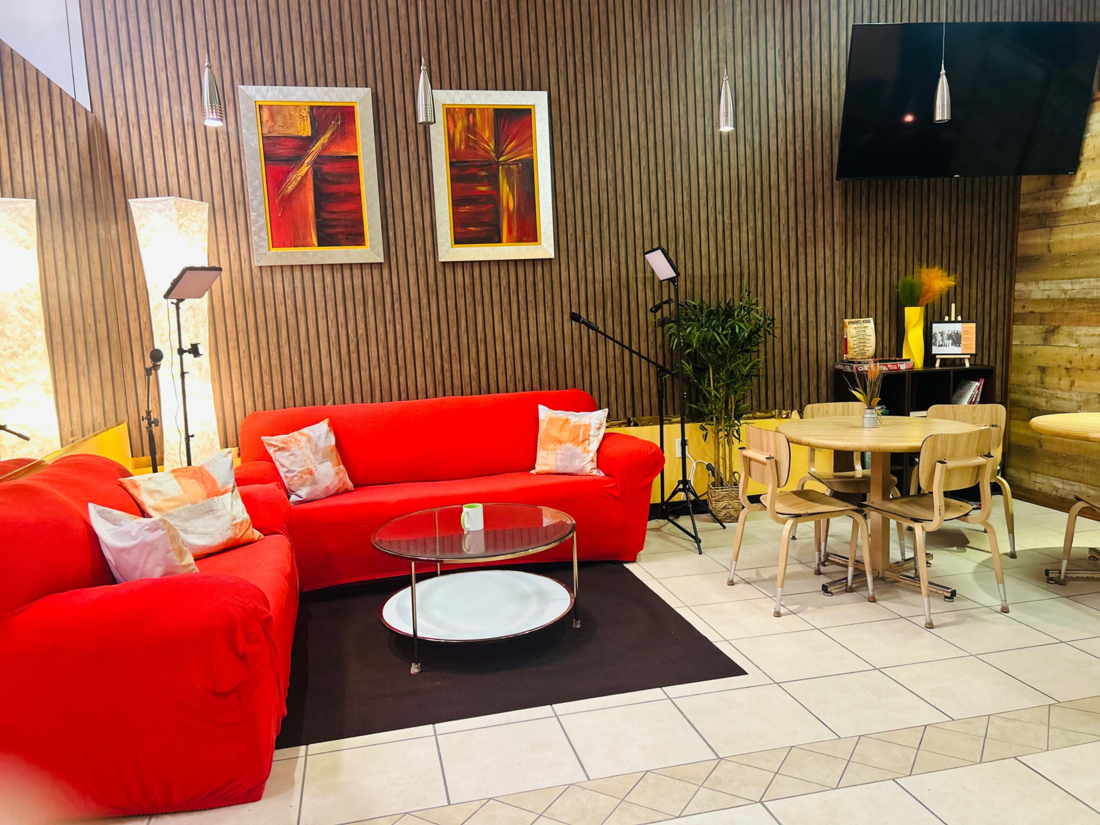 A brightly colored living room features a red couch, a circular coffee table, wooden dining chairs, and wall-mounted artwork.