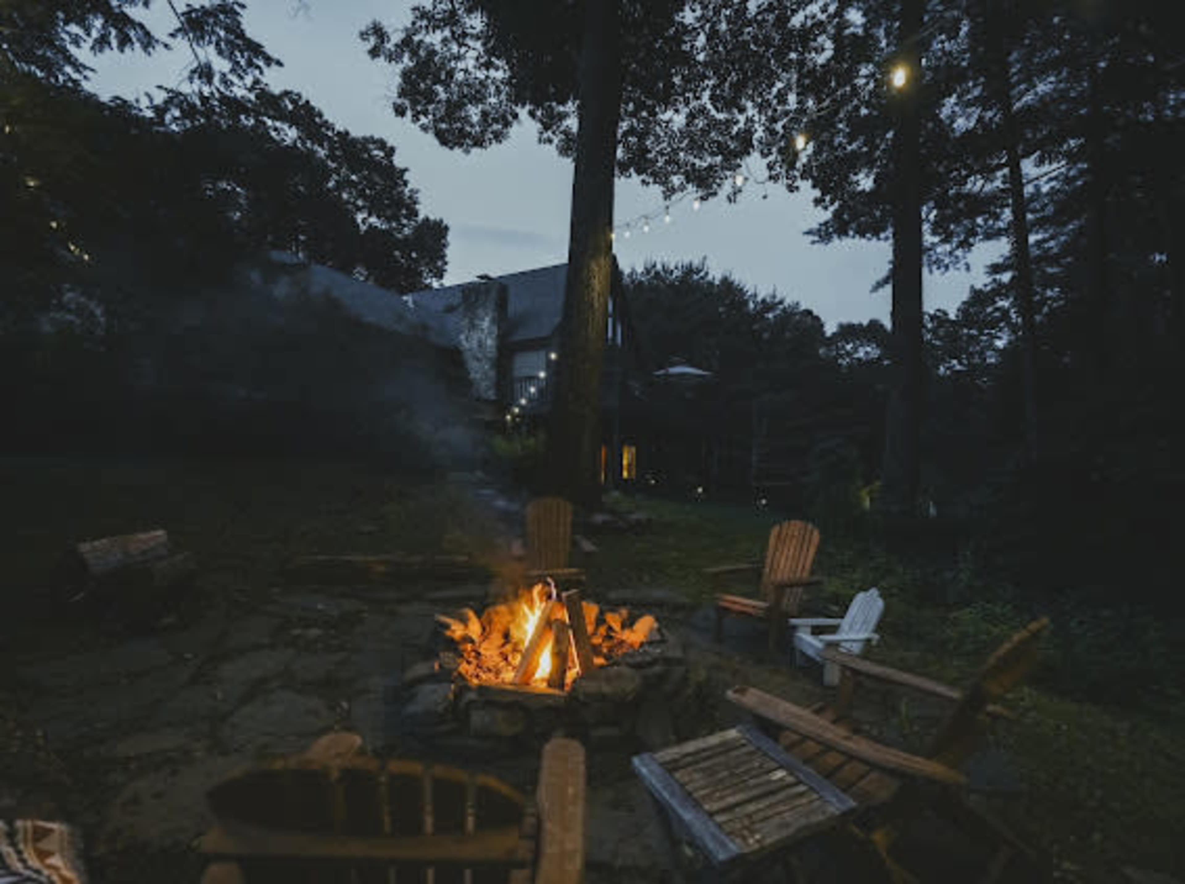 A fire sits at the center of a clearing, surrounded by wooden chairs, with a house partially visible in the background under a twilight sky.