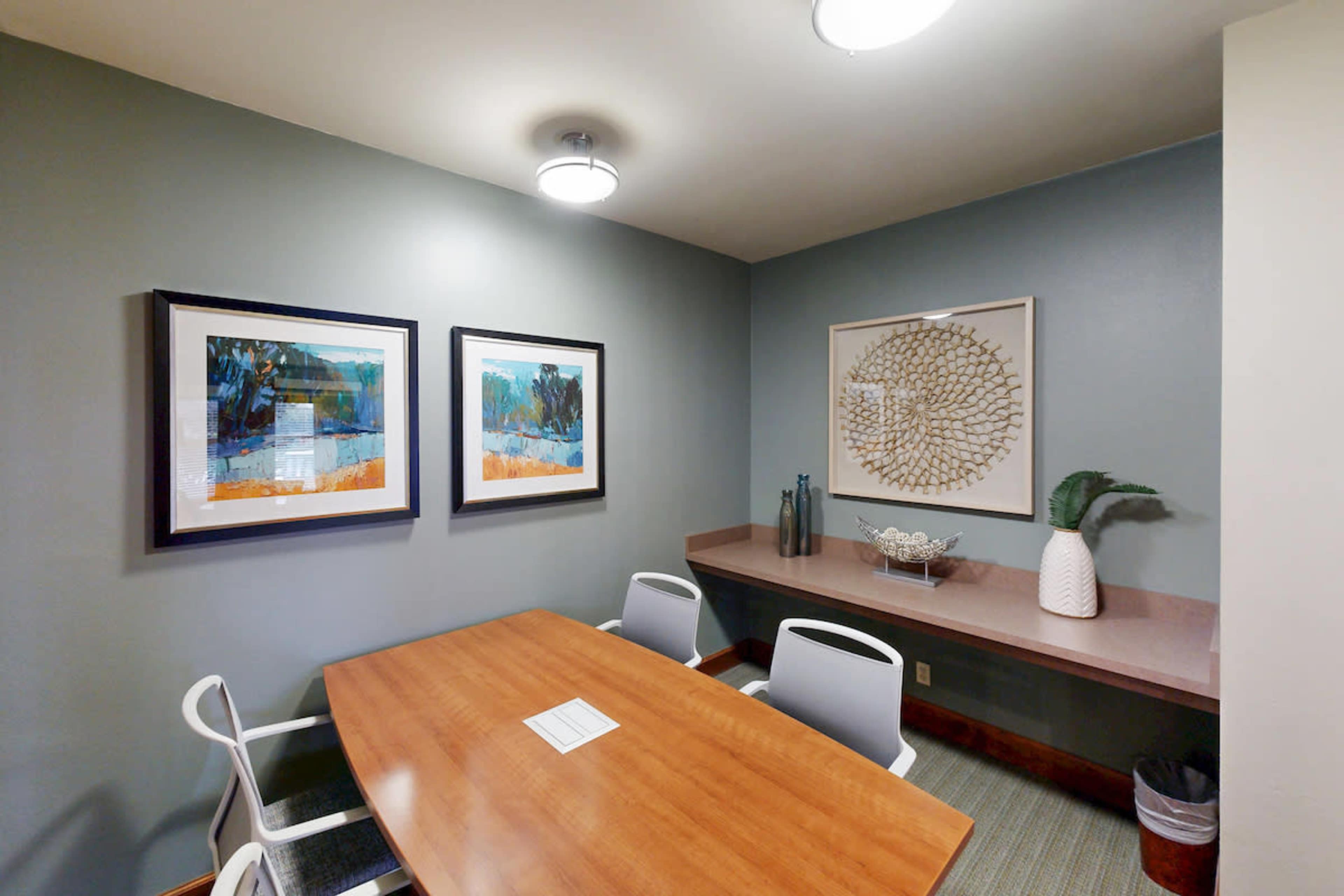 The image shows a small meeting room with a wooden table surrounded by four chairs, decorated with framed artwork and a wall sculpture.