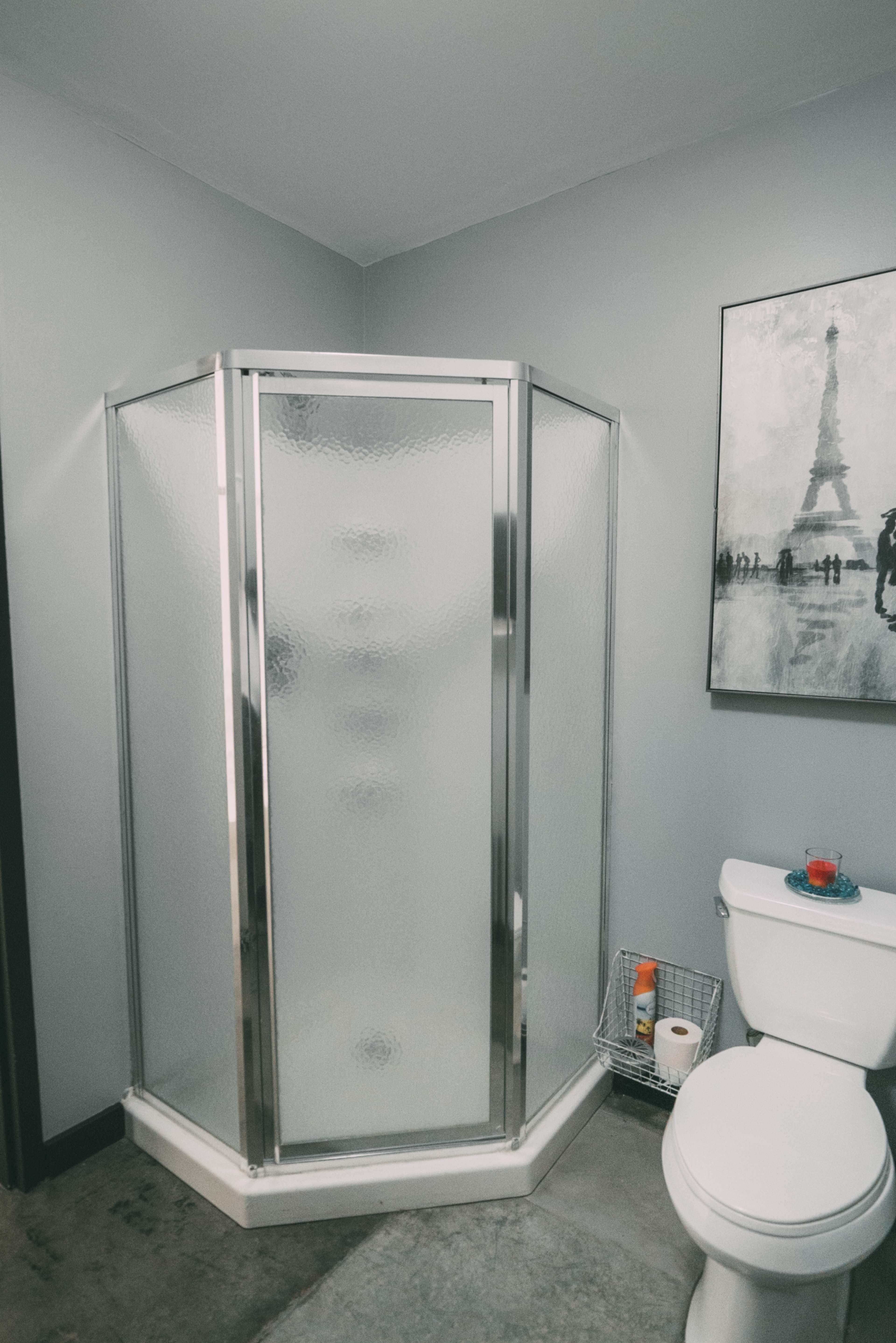 A corner shower stall with a glass door is positioned next to a toilet in a bathroom featuring gray walls and a framed picture of the Eiffel Tower.