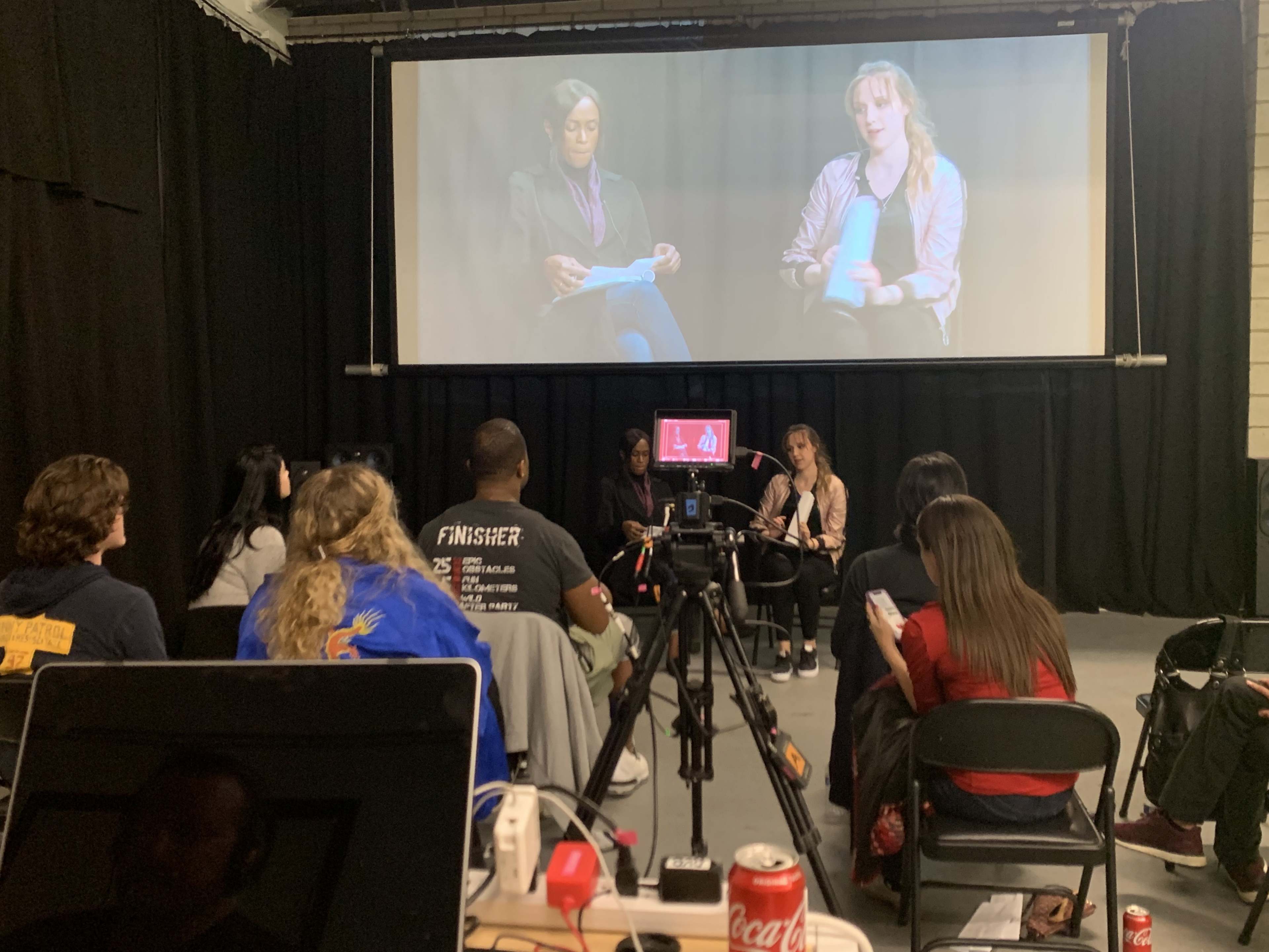 A group of audience members watches a discussion on a large screen featuring two speakers in a studio setting.