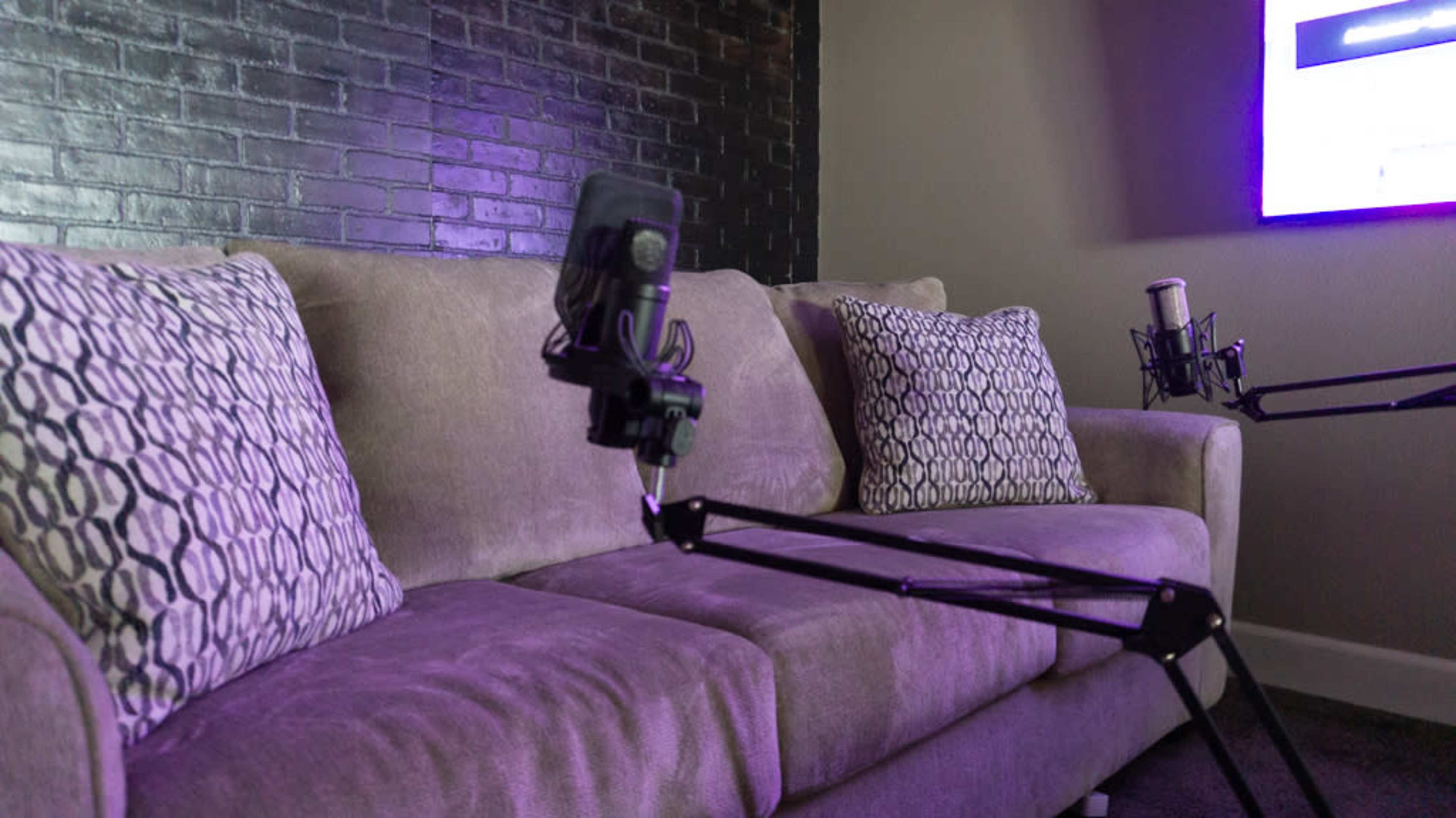 The image shows a beige couch with two patterned pillows and two microphones mounted on stands positioned in front of it.
