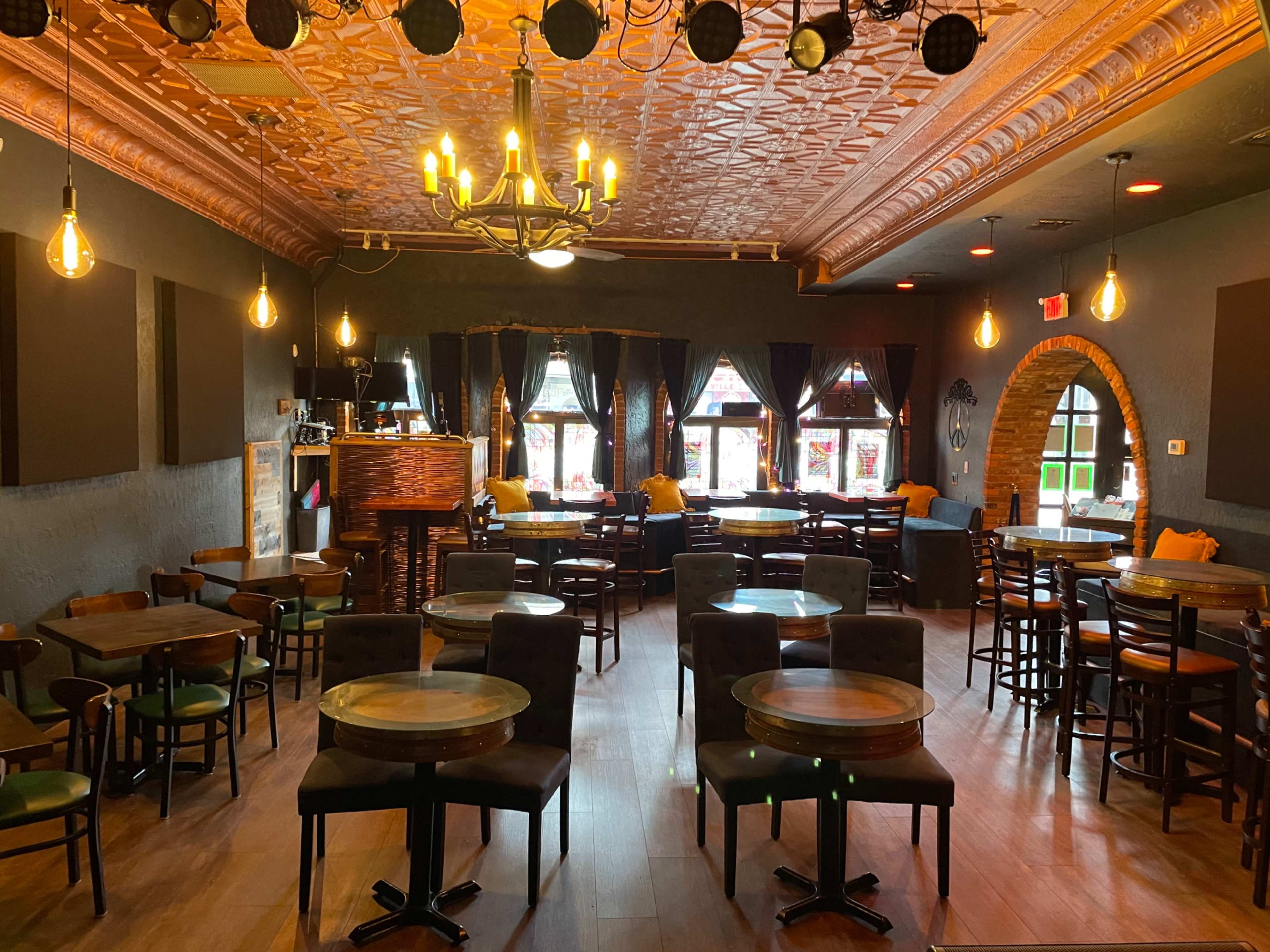 A dimly lit bar area with wooden tables and chairs, adorned with chandeliers and vintage decor, featuring large windows.