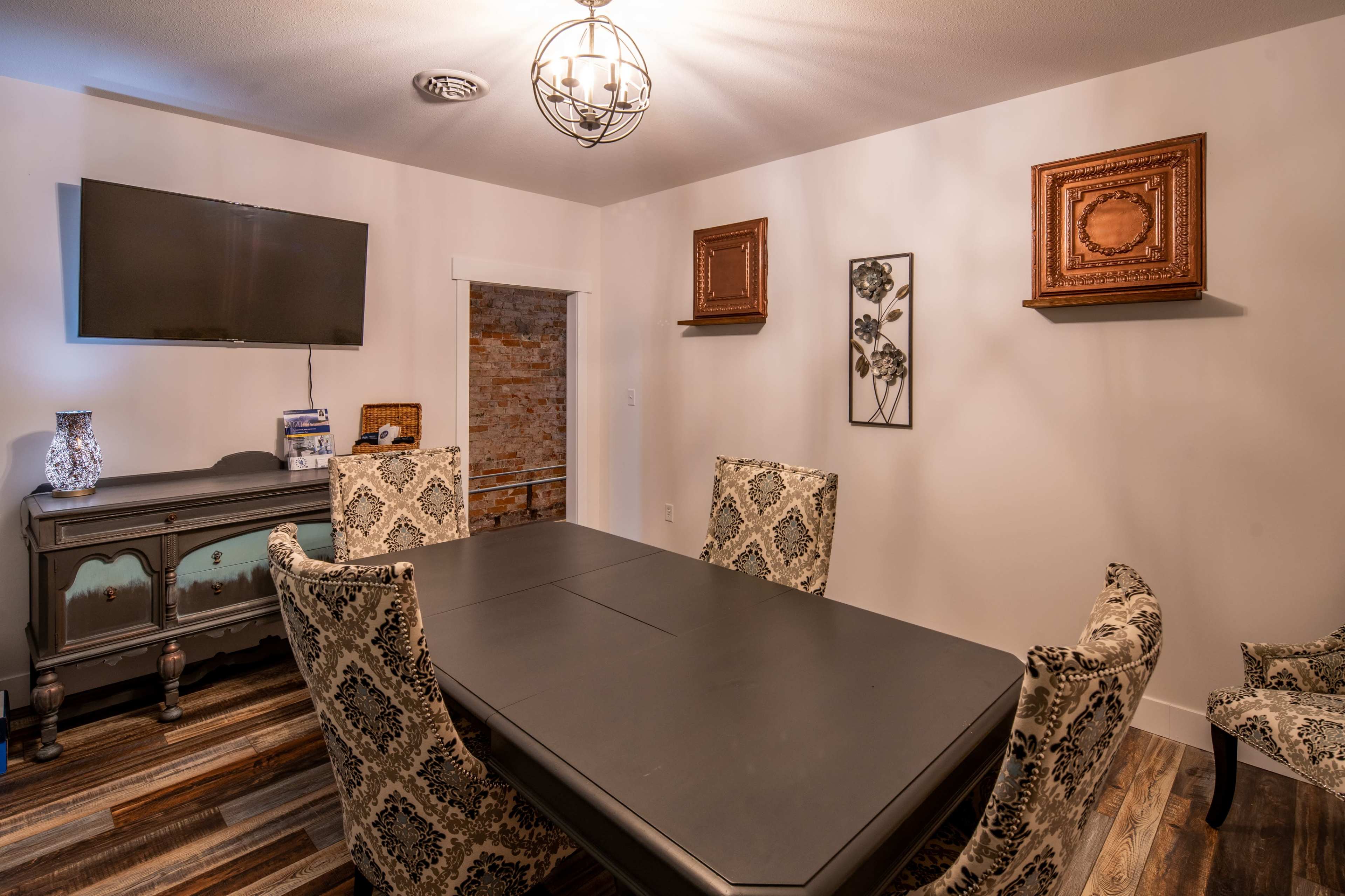A dining room with a large black table surrounded by ornate chairs, a television mounted on the wall, and decorative elements including framed art and a cabinet.