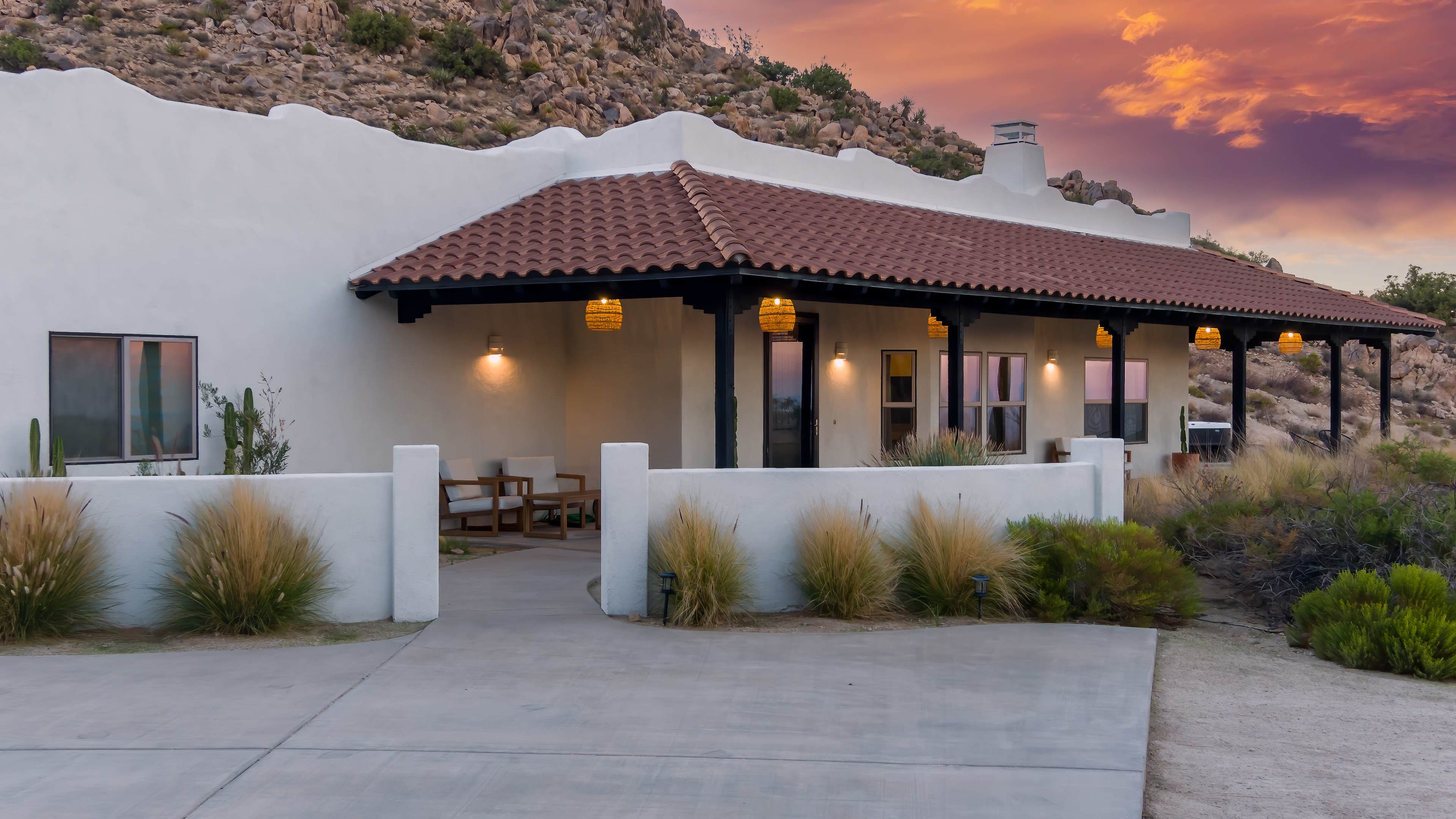 A single-story house with a tiled roof and white exterior sits against a rocky hillside under a colorful sunset sky.