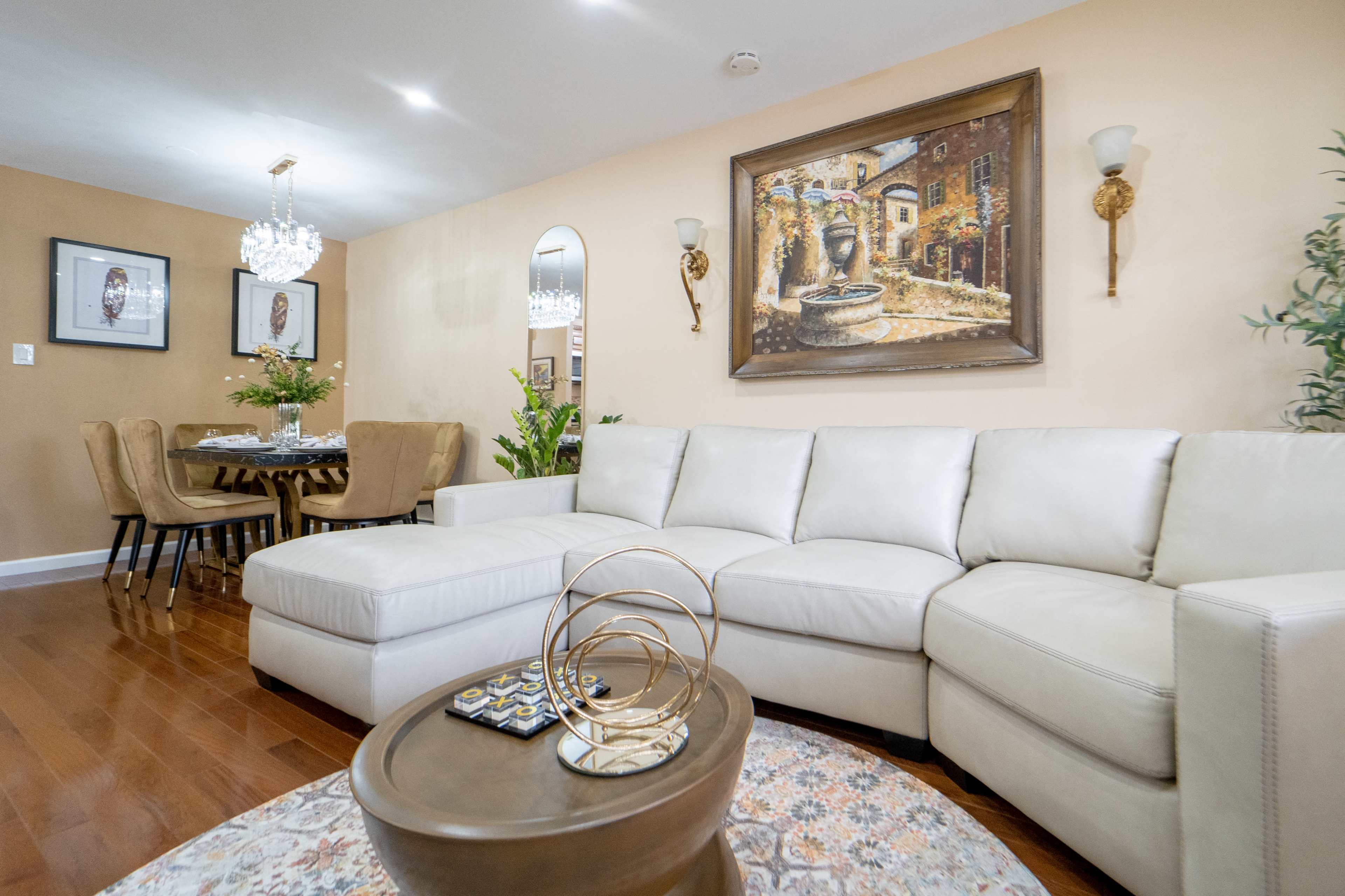 The living room features a cream-colored sectional sofa, a circular wooden dining table with upholstered chairs, and decorative artwork on the walls.
