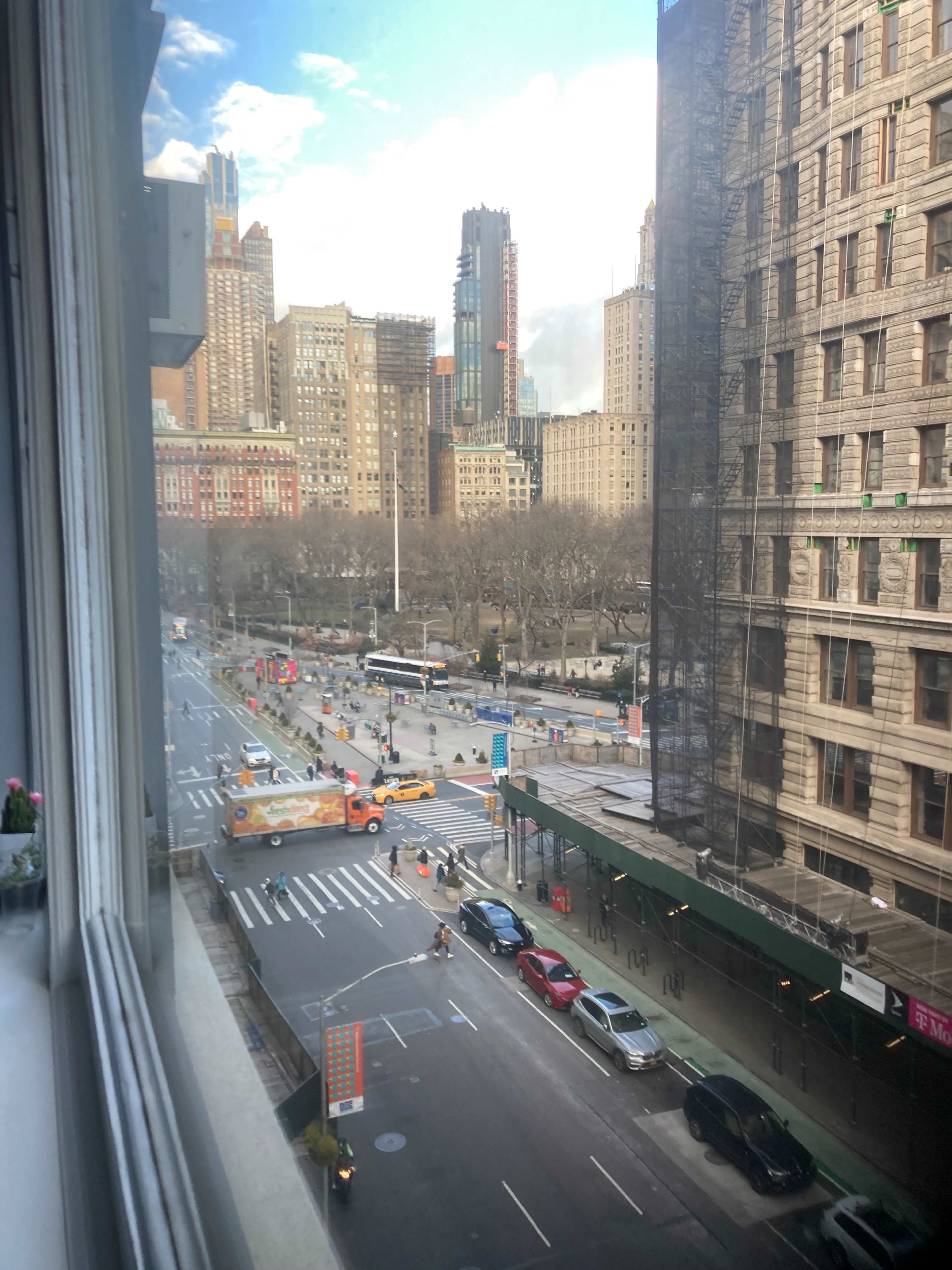 A view of a busy city street with vehicles, buildings, and a park in the background, observed from a window.