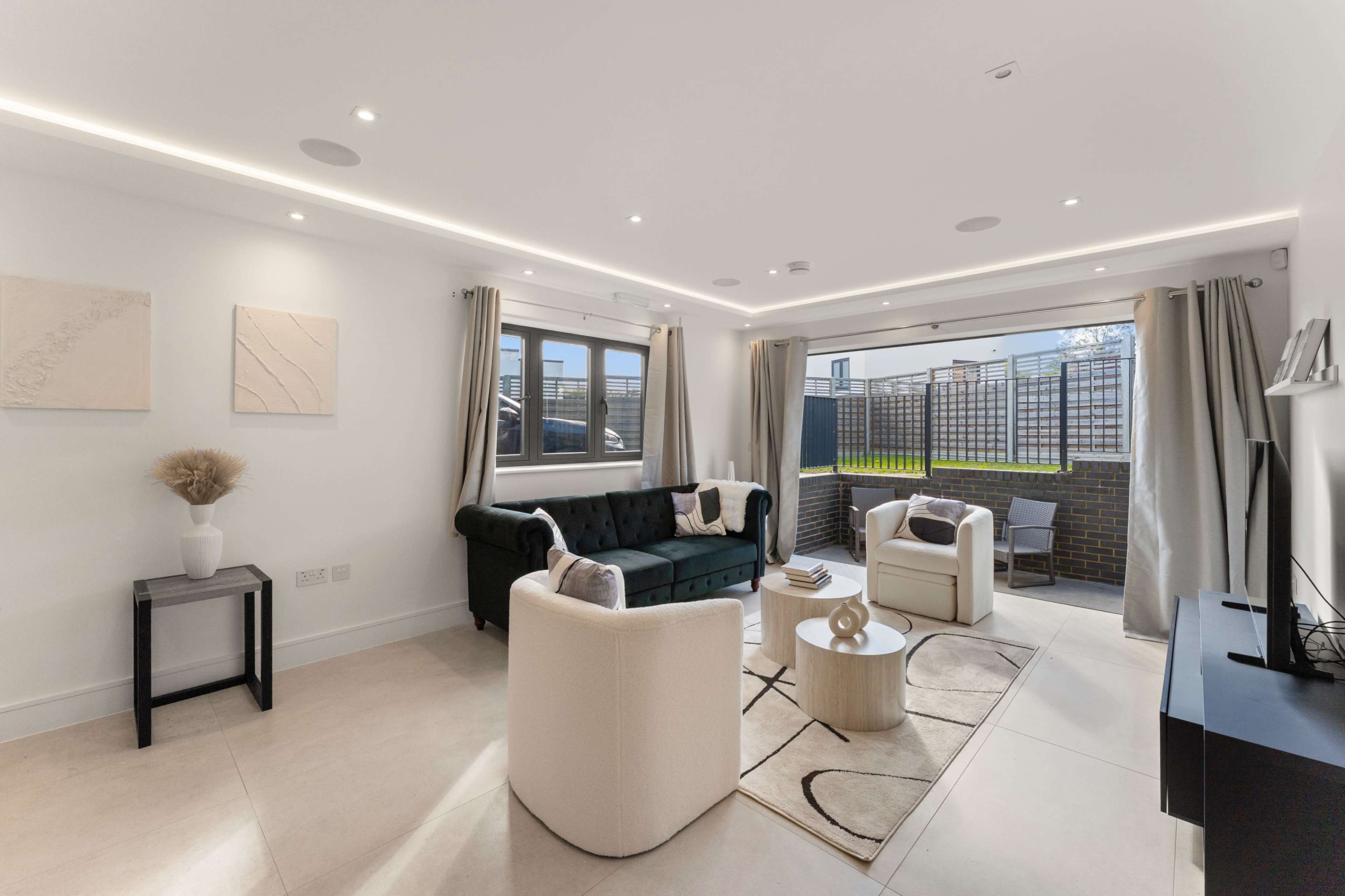 The image shows a modern living room with a green sofa, two white armchairs, and large windows that open to an outdoor space, complemented by neutral-colored decor and a round coffee table.