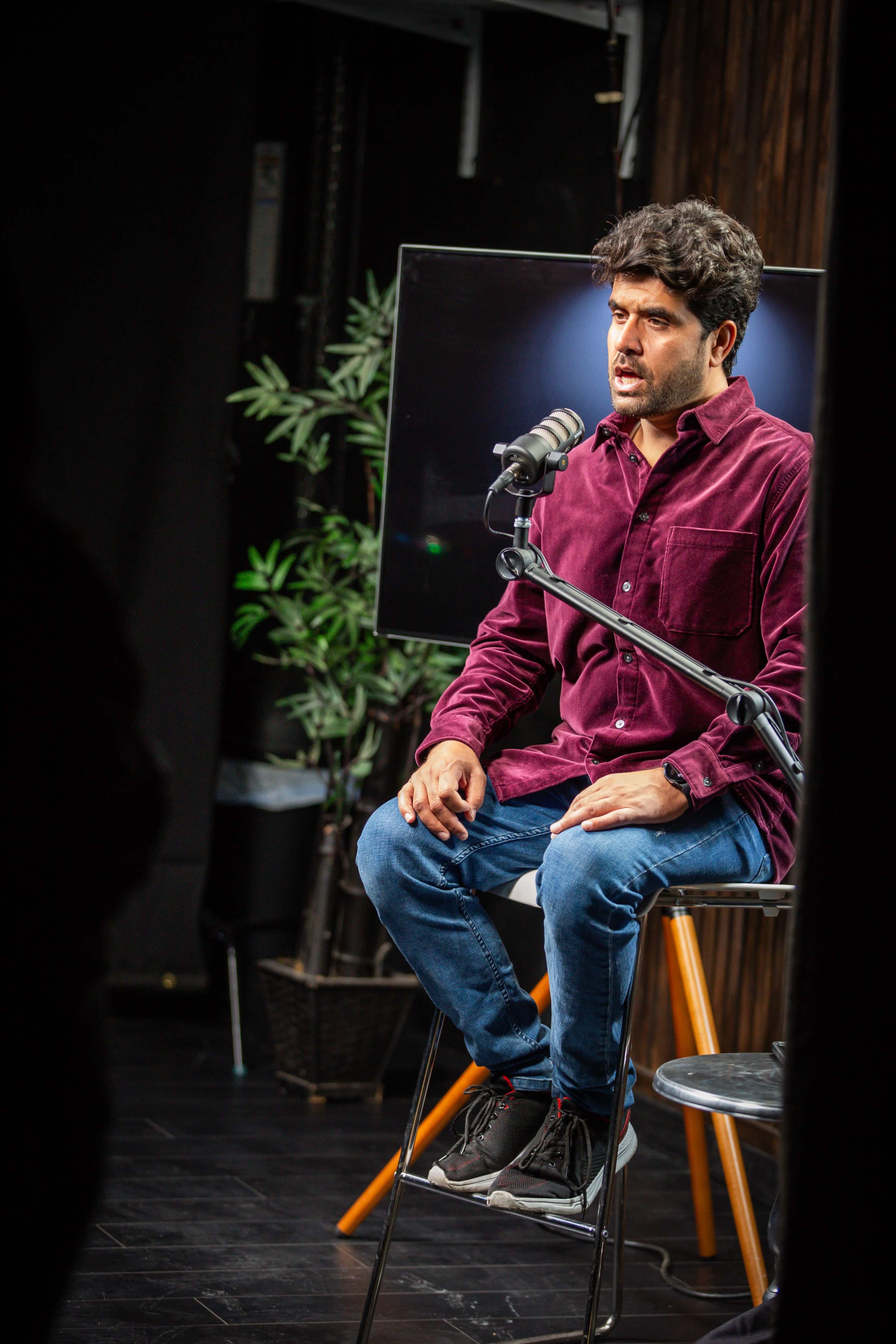 A man with dark hair sits on a stool in front of a microphone and a television screen while wearing a burgundy shirt and jeans.