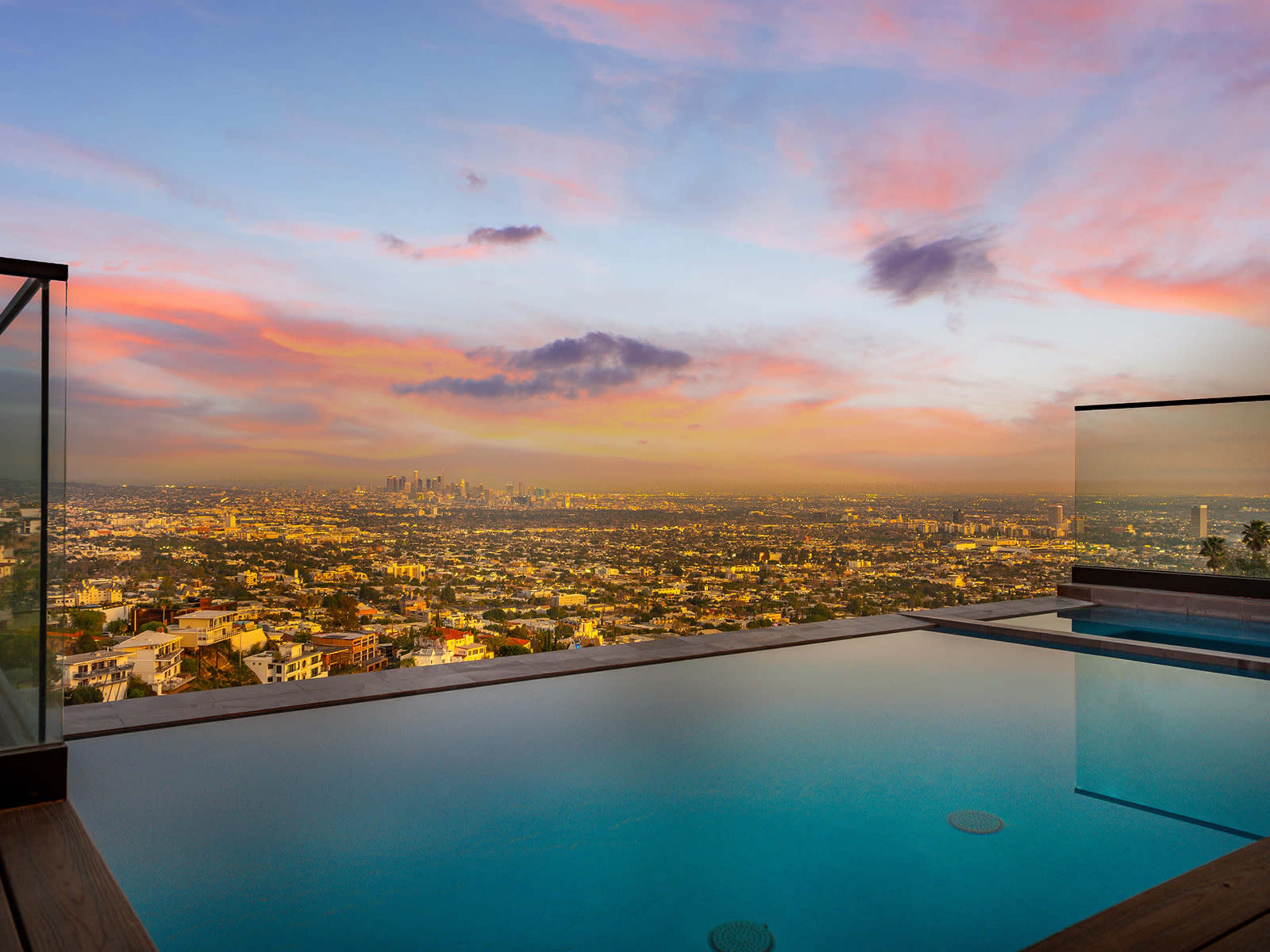 A panoramic view of Los Angeles is visible from a modern poolside, featuring a clear swimming pool with a backdrop of a colorful sunset sky.