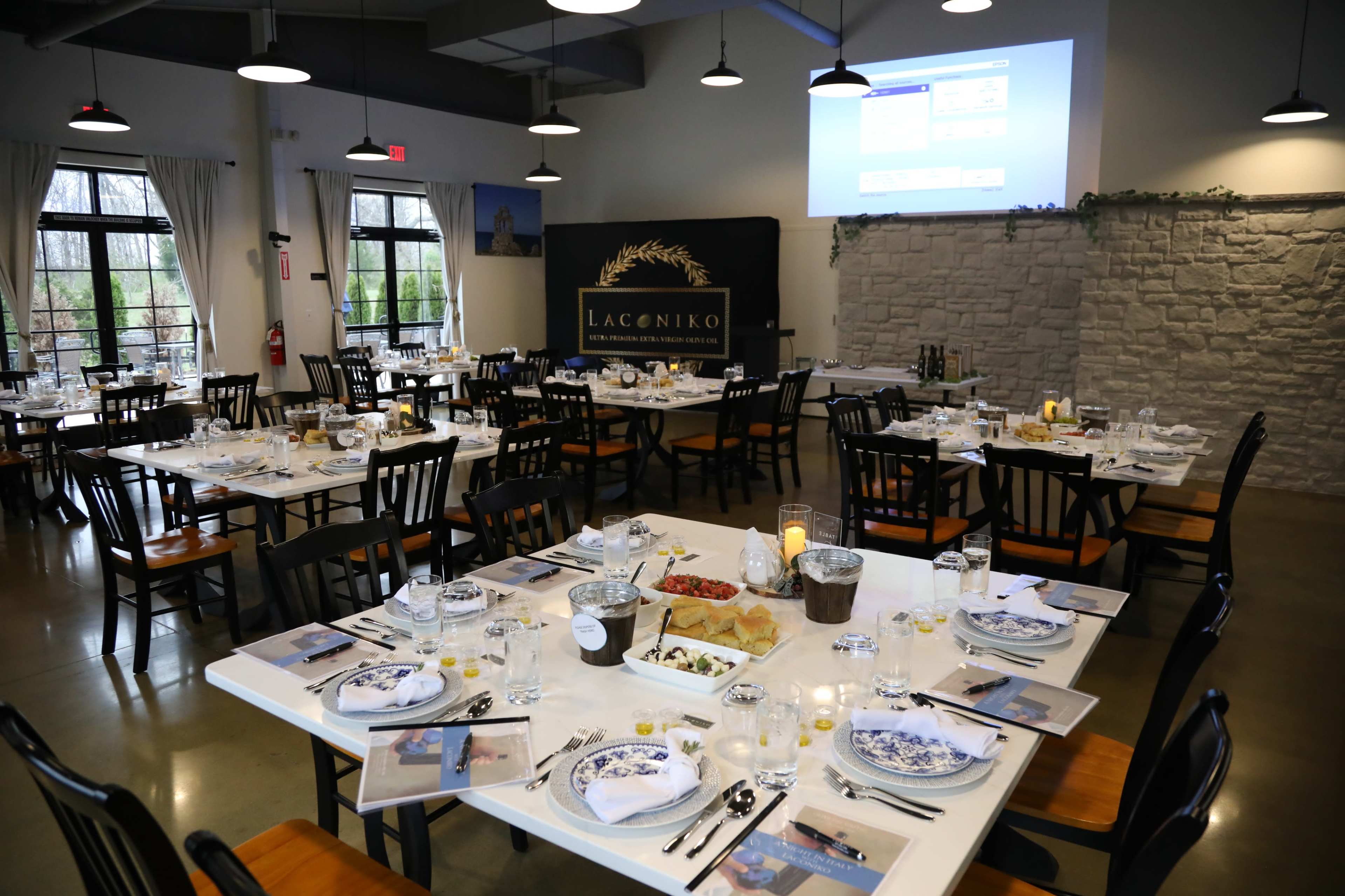 The image shows a dining room set with multiple tables arranged for a meal, featuring elegant table settings and a large projector screen on the wall.