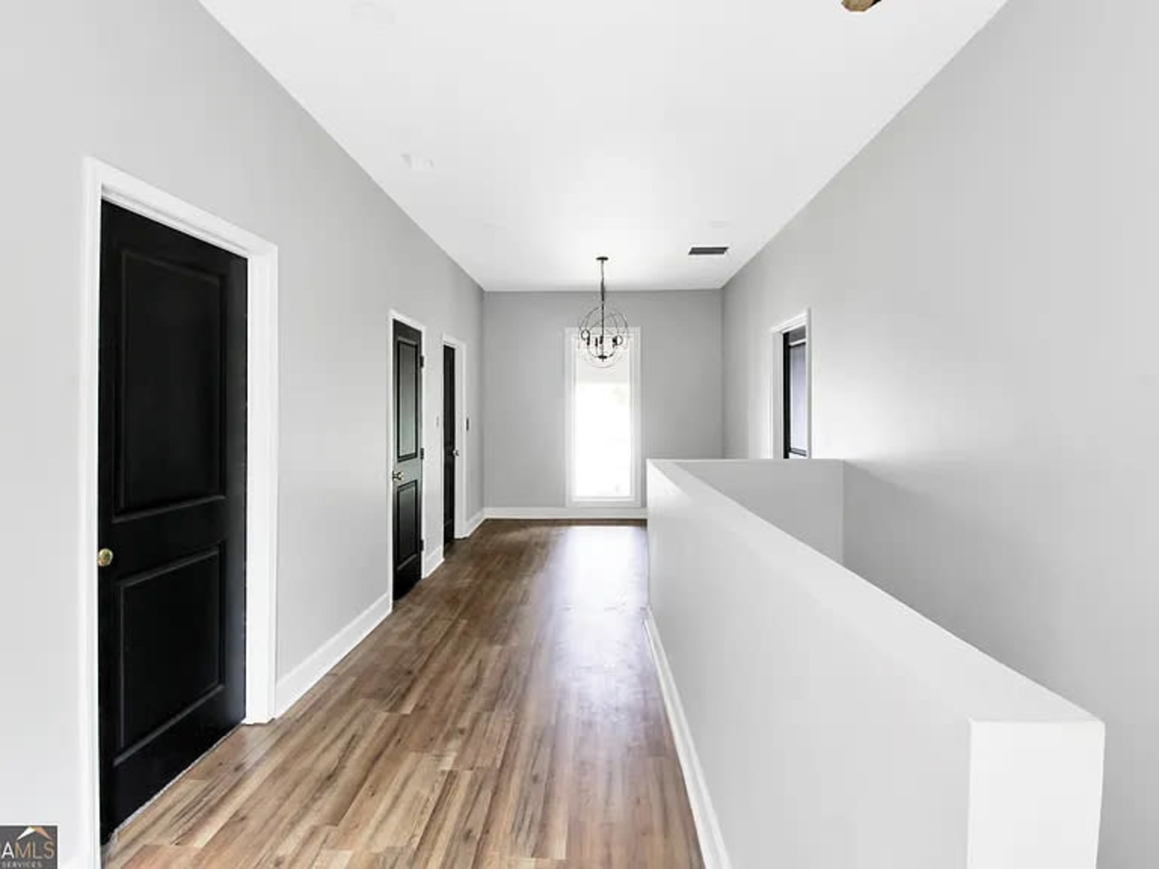 A hallway with light gray walls, black doors on either side, and a wooden floor leads to a window at the end.
