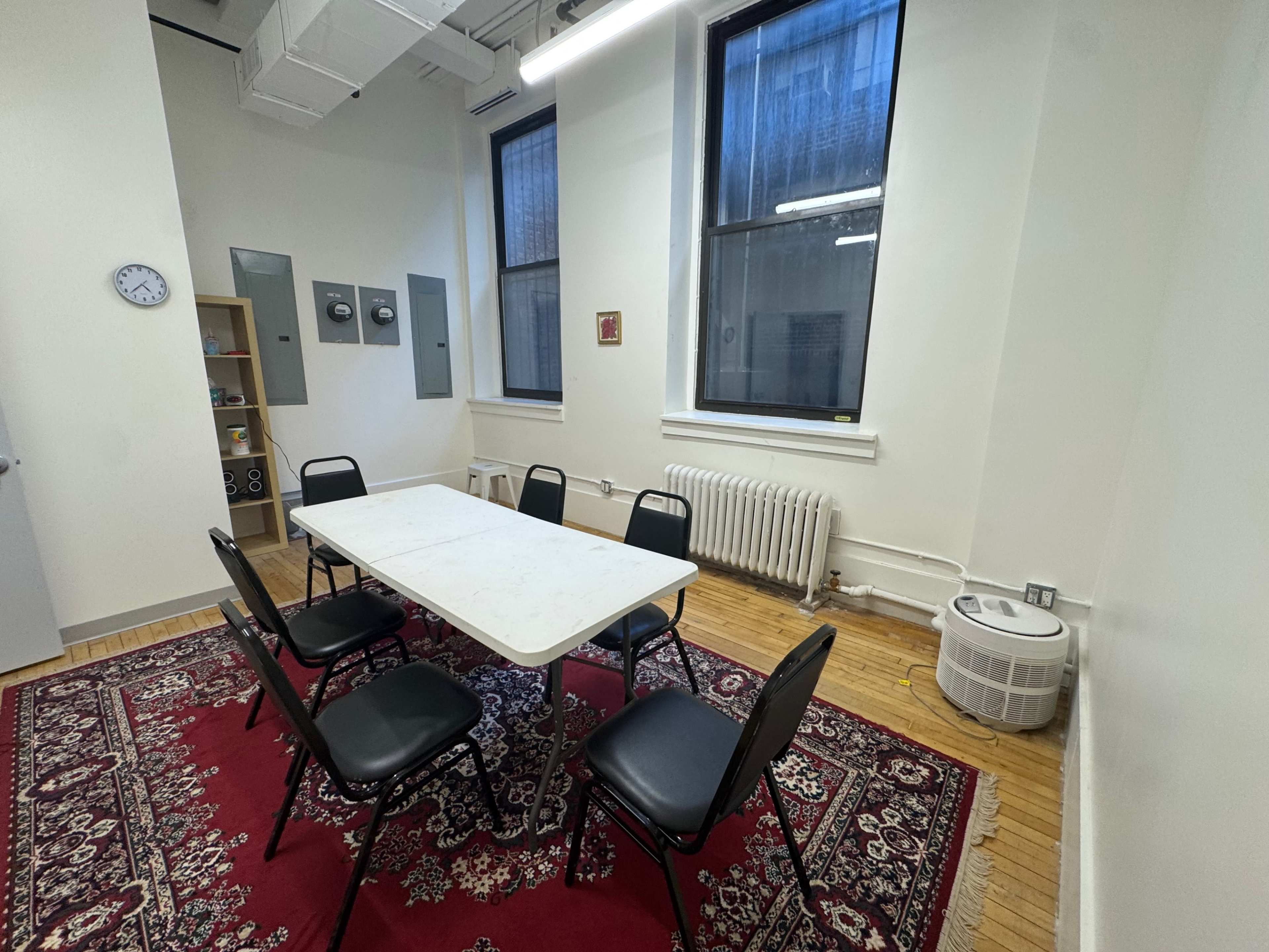 The image shows a simple meeting room with a rectangular table surrounded by six black chairs, on a red rug, with windows, a clock, and heating units in view.