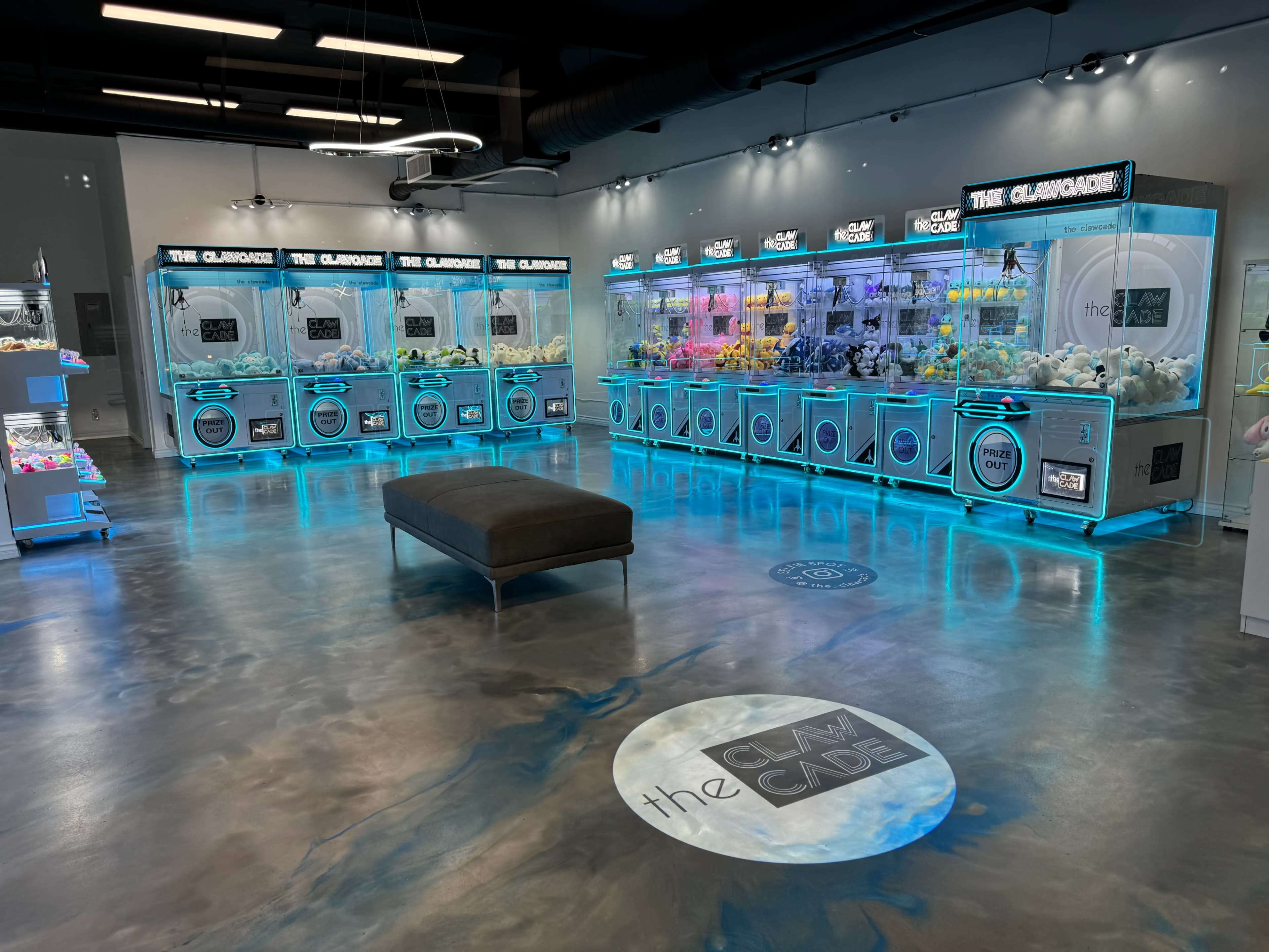 The image shows an arcade resembling a claw machine area with brightly illuminated displays, featuring multiple claw machines and a central bench in a modern, spacious setting.