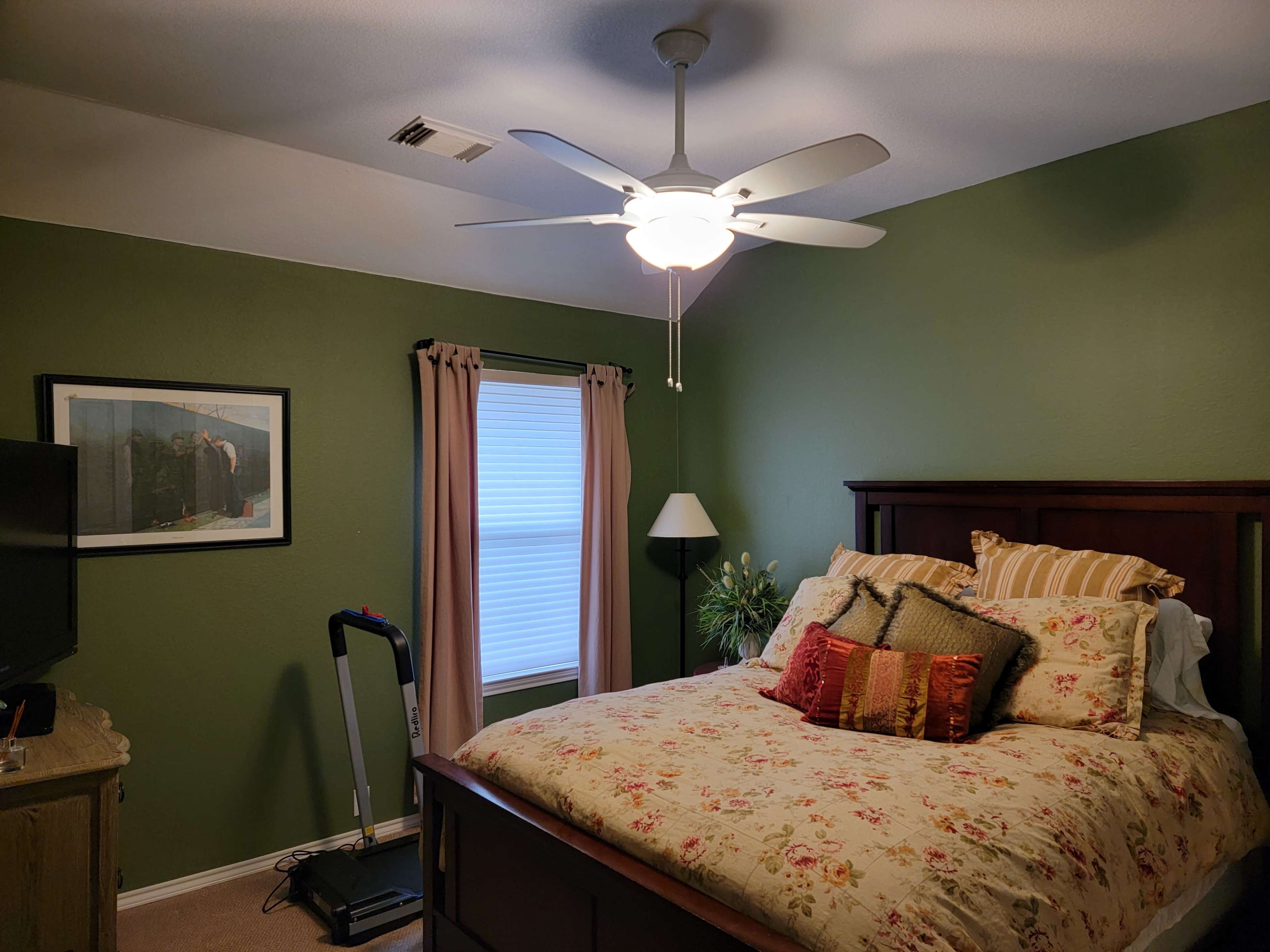 The image shows a bedroom with a green wall, a bed covered in floral bedding, a ceiling fan, and a window with light curtains.