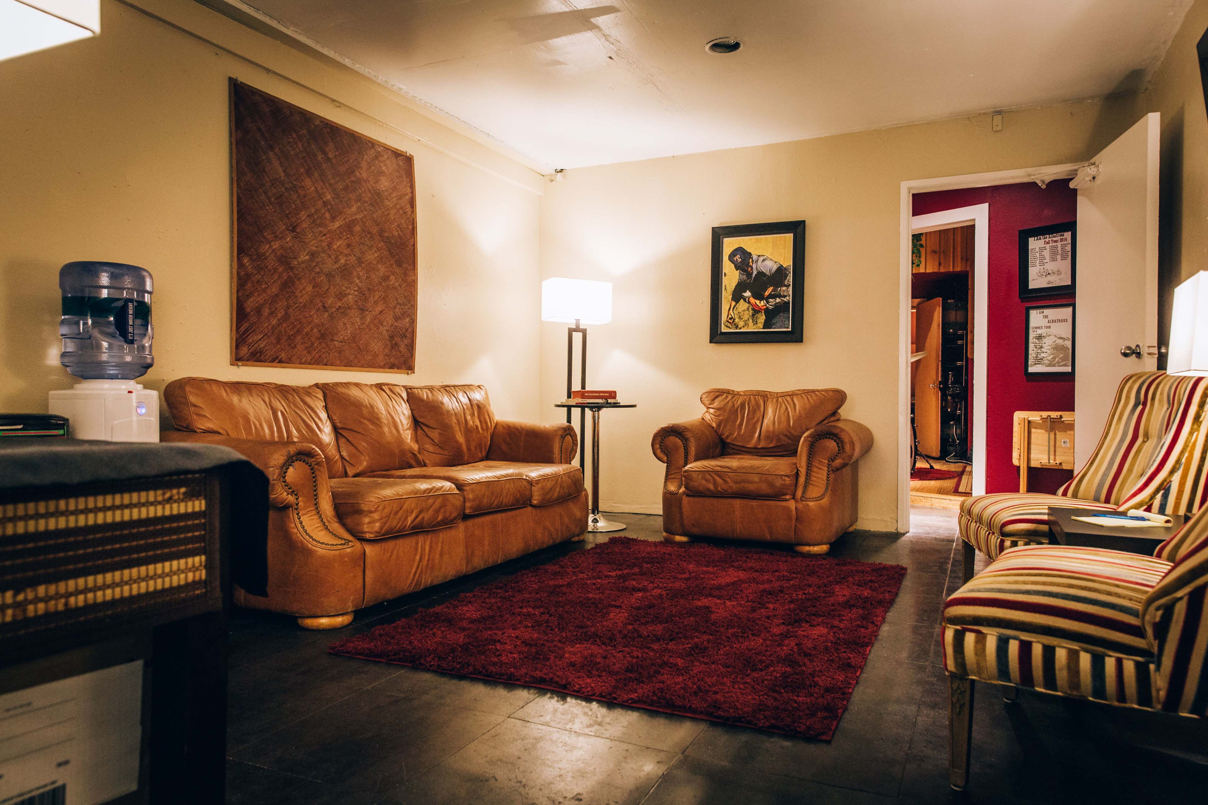 A cozy living room features two leather sofas, a striped armchair, a lamp, and a water cooler, with a red rug on a dark tiled floor.