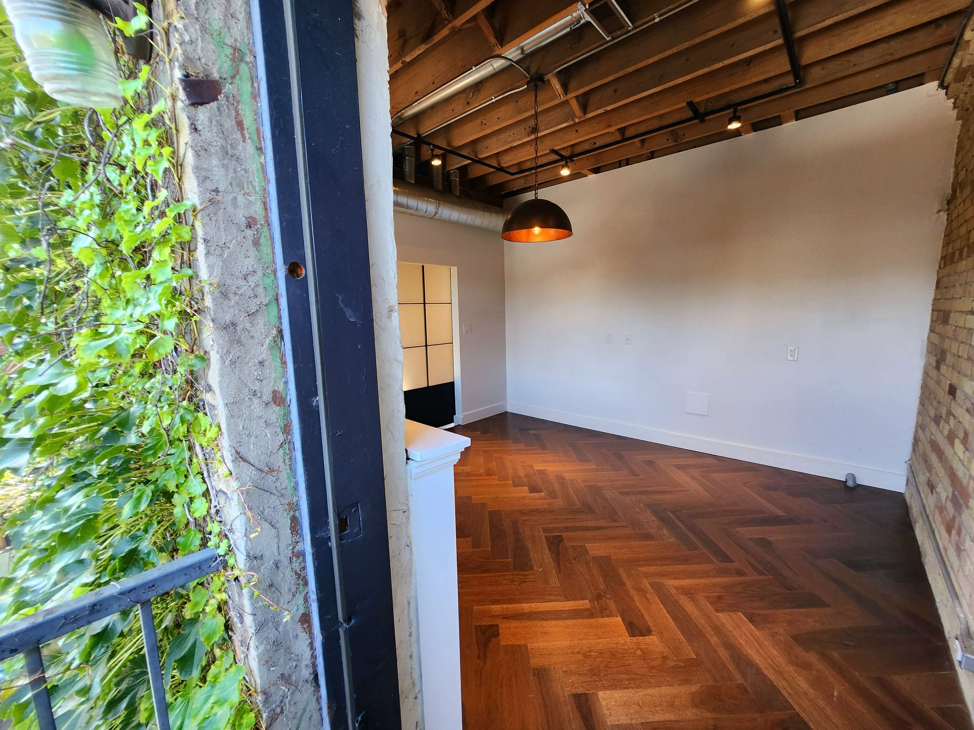 A room with wooden flooring, exposed beams, and a large window opening to a balcony surrounded by greenery.