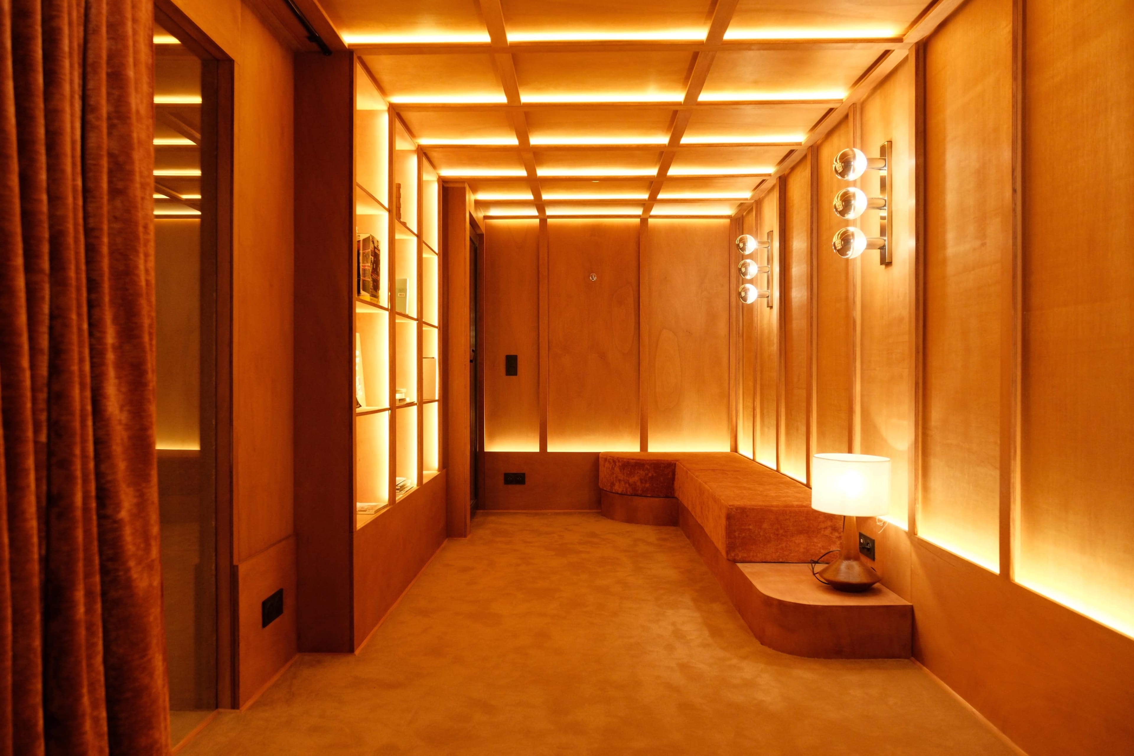 The image shows a warm-toned interior space featuring wooden paneling, recessed lighting, and a small seating area with a lamp.