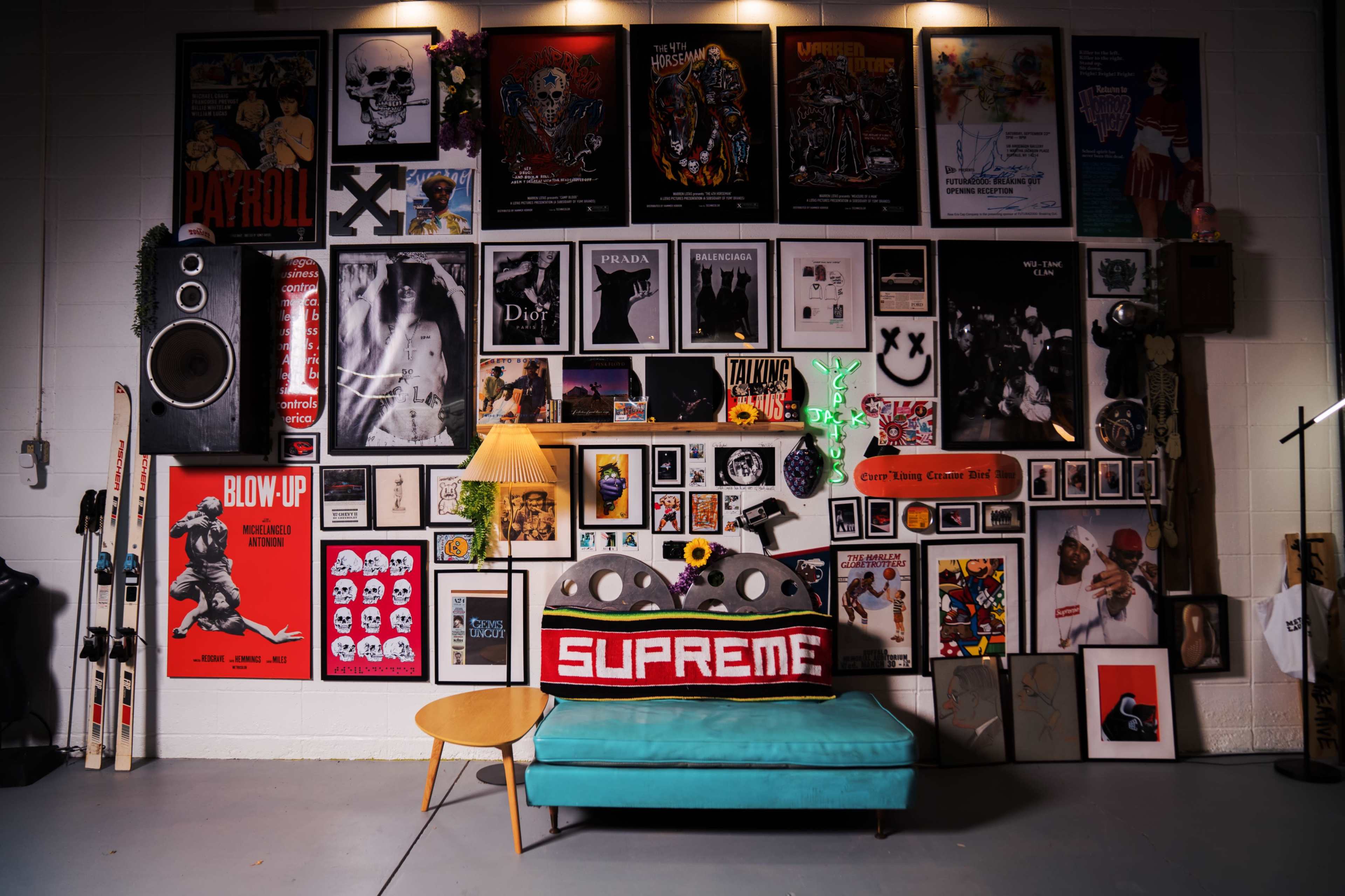 The image features a colorful gallery wall adorned with various framed posters and artwork, above a teal sofa with a red Supreme pillow and a lamp, surrounded by decorative items.