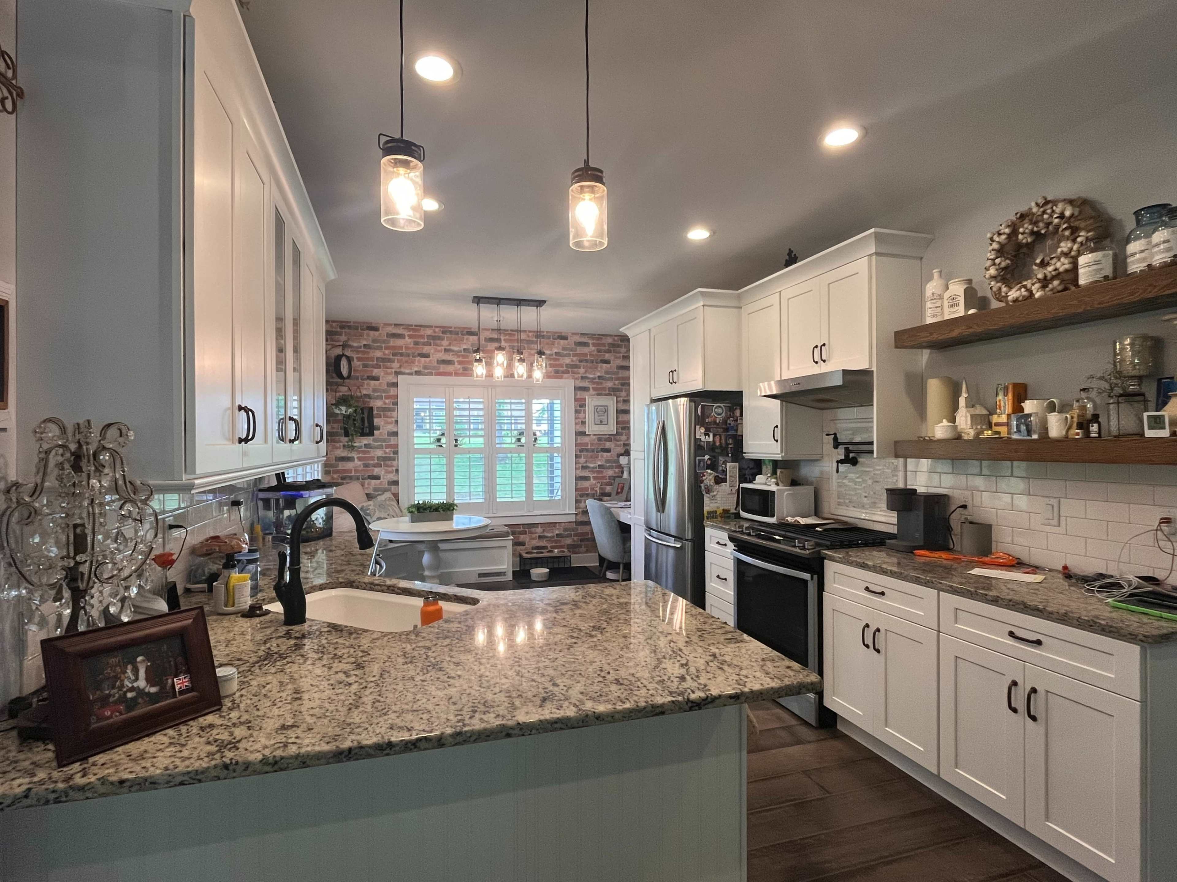 A modern kitchen features white cabinetry, granite countertops, and a mix of pendant and recessed lighting above an island, with a view of a brick-patterned wall and a window.