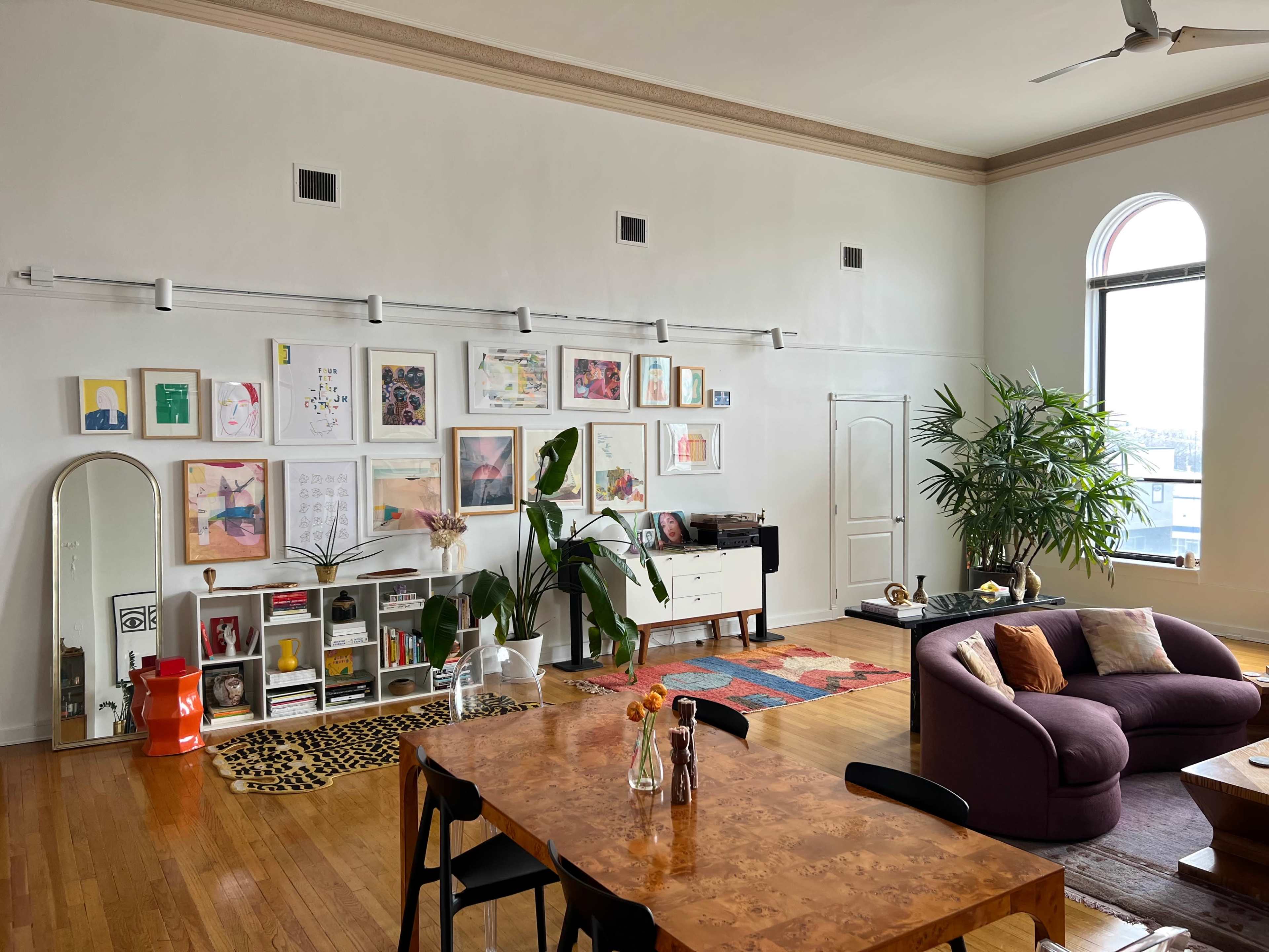 The image shows a modern living room with a purple sofa, a large dining table, a gallery wall of framed art, and a variety of plants.