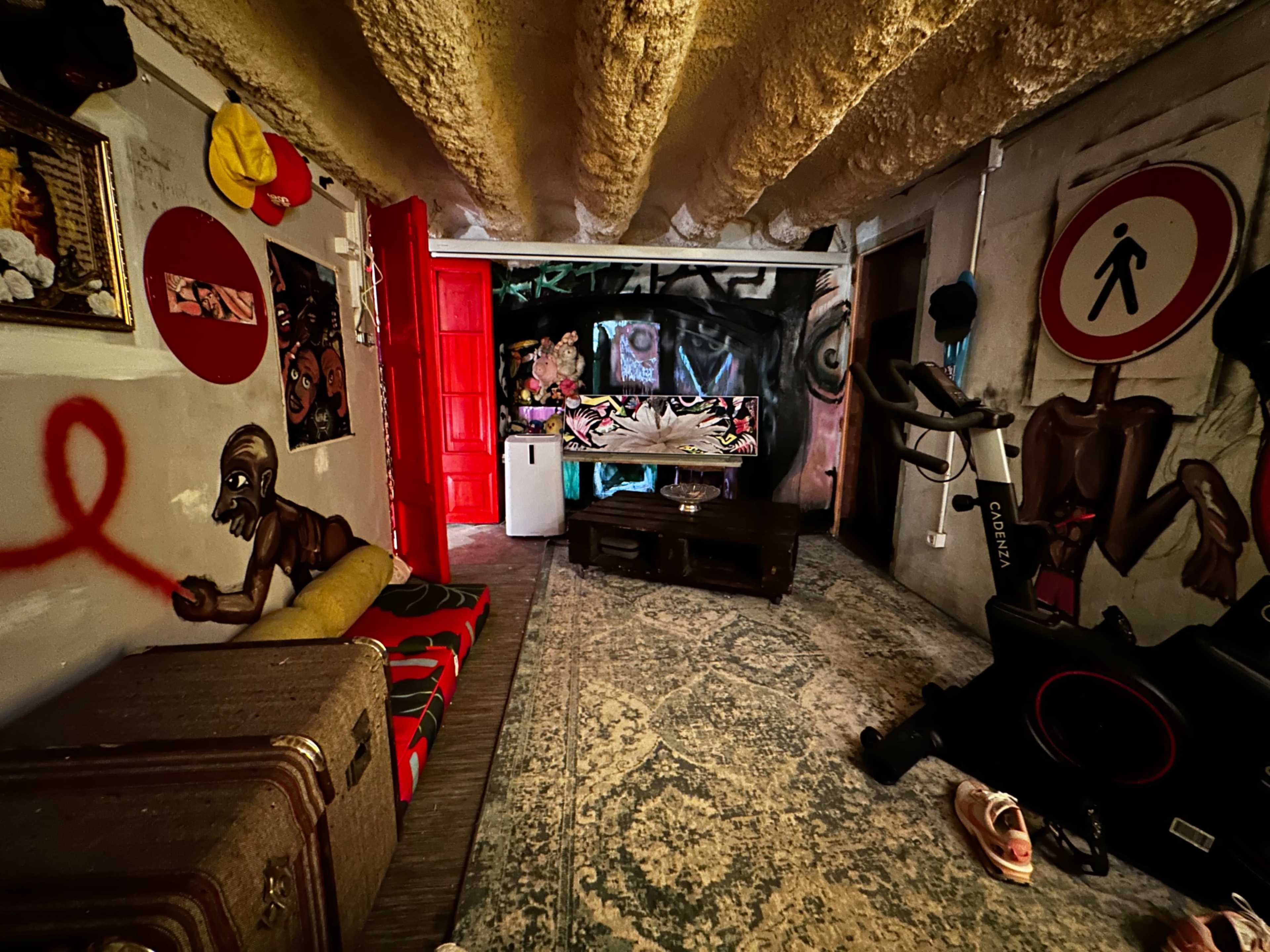 The image depicts a basement room featuring colorful graffiti art, a red door, a workout bike, and various furniture pieces arranged around a large area rug.