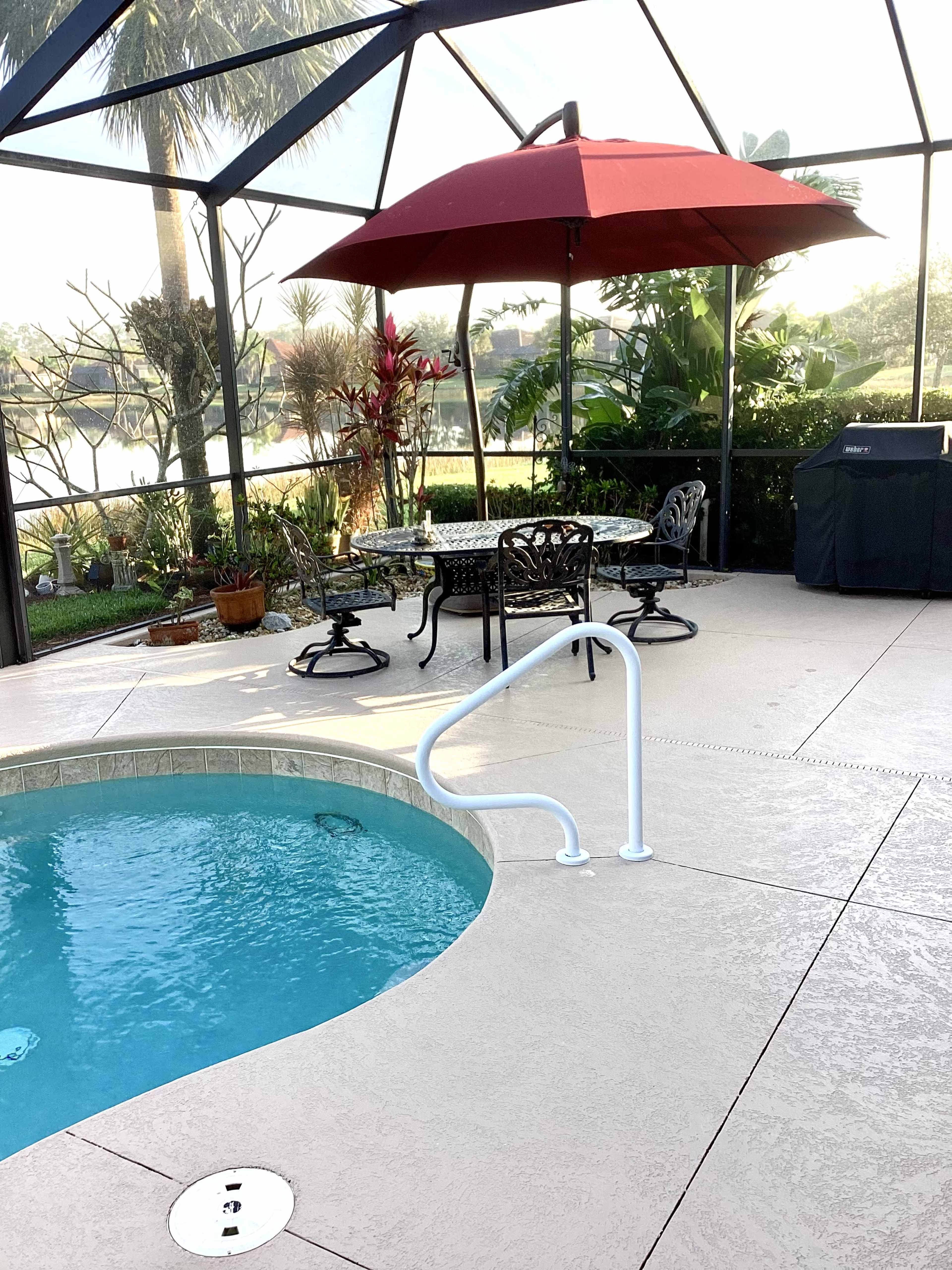 A patio area features a swimming pool, a round table with chairs under a red umbrella, and tropical plants in a screened enclosure.