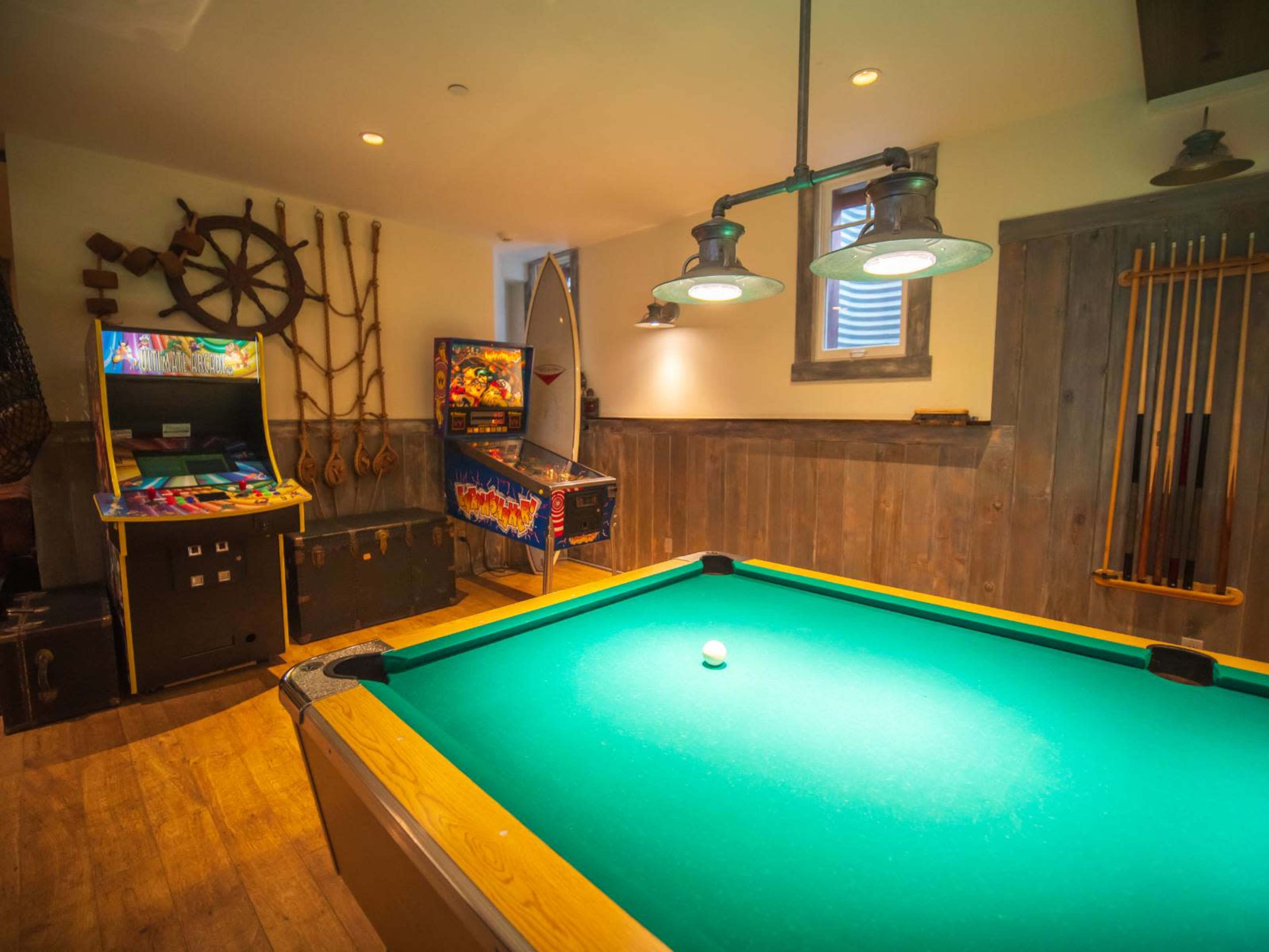 The image shows a game room featuring a pool table, arcade machines, and wall-mounted pegboards with sports equipment.