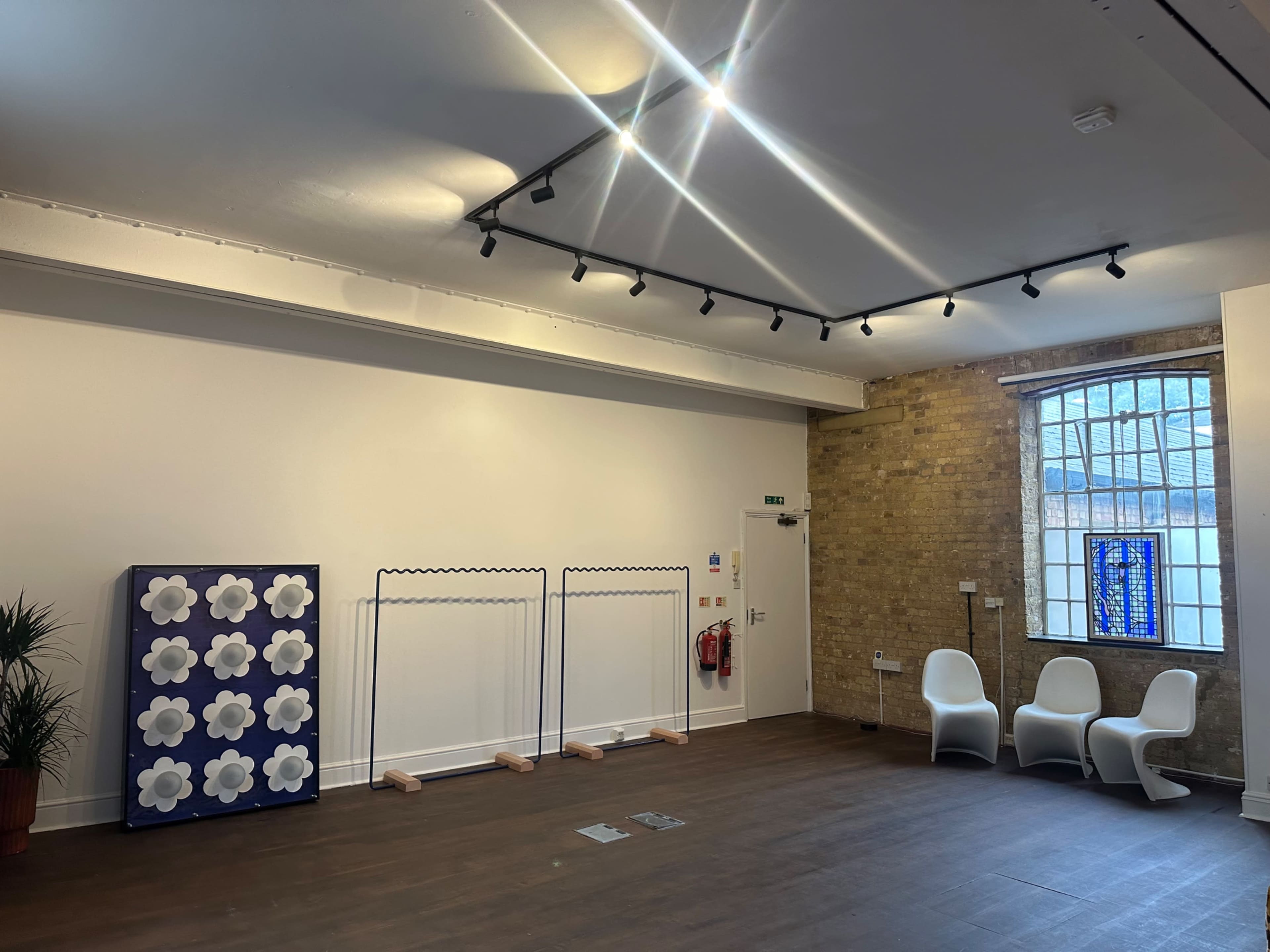 The image shows an empty room with wooden flooring, equipped with a large decorative partition featuring flowers, two clothing racks, a plant, and three white chairs against a brick wall.