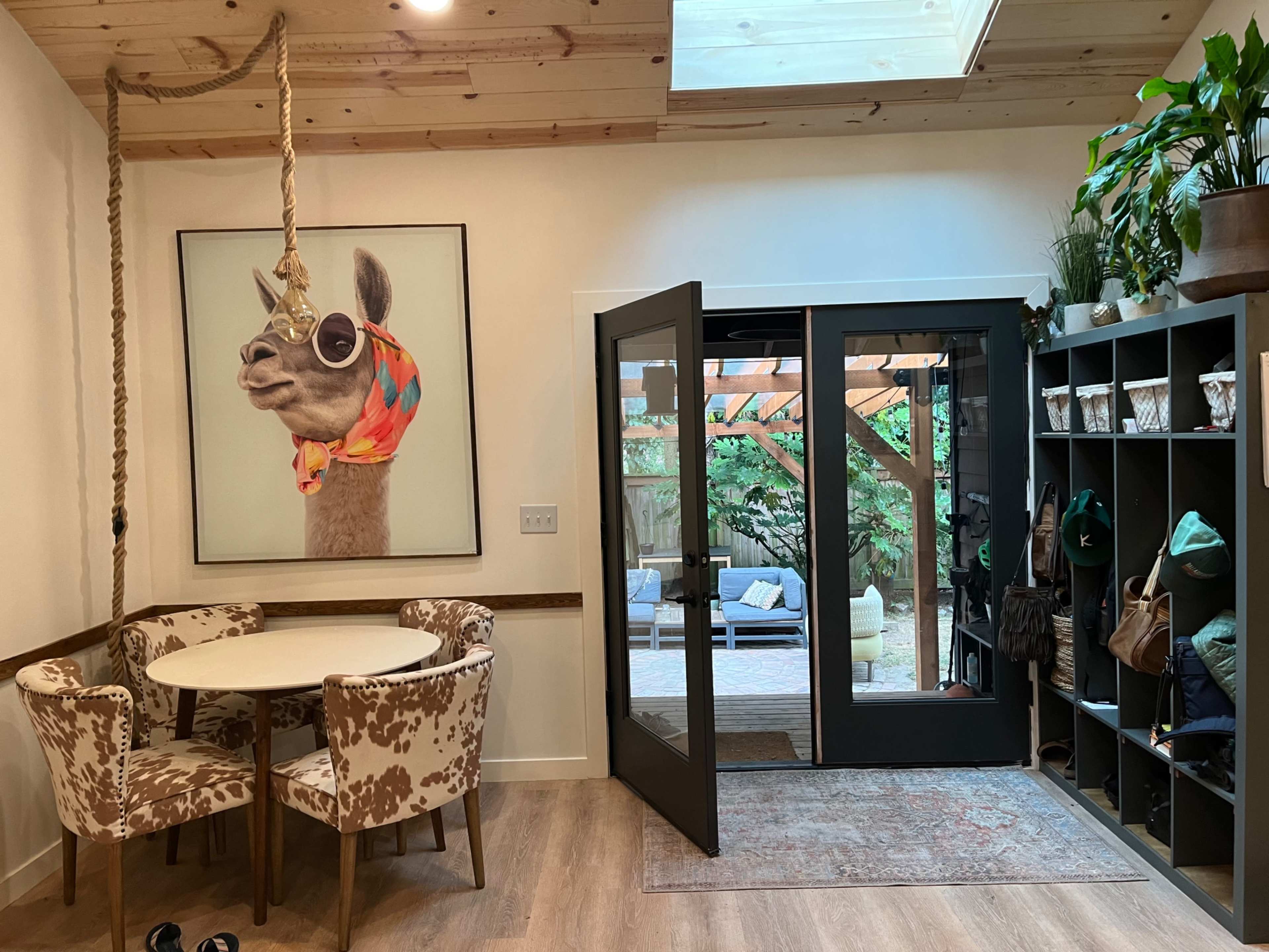 An entryway with a circular table and patterned chairs, a large framed portrait of a llama wearing sunglasses, and a pair of glass doors leading to a patio area.