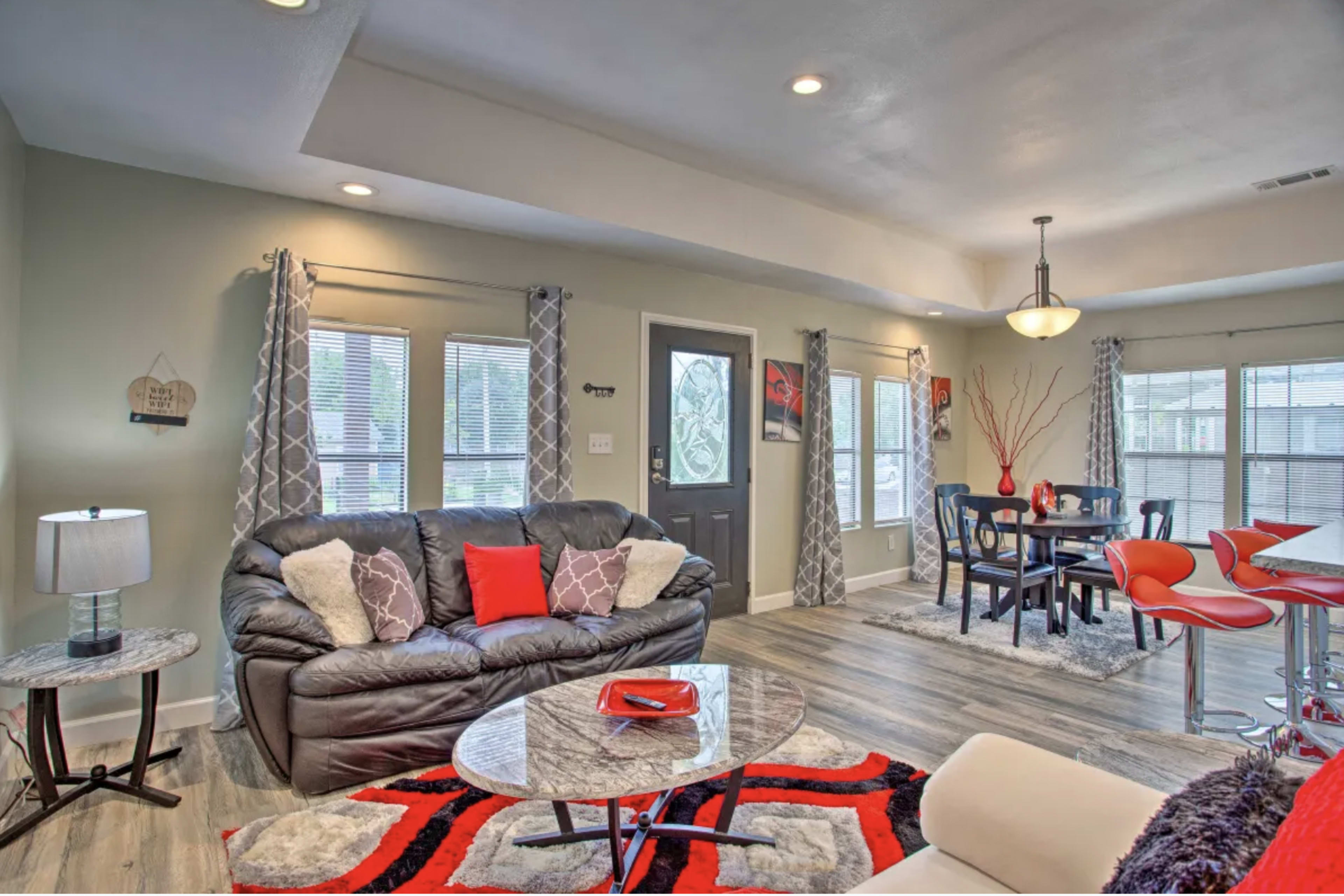 A modern living room and dining area with a black leather sofa, a round coffee table, and a dining table surrounded by red chairs, illuminated by natural light through multiple windows.