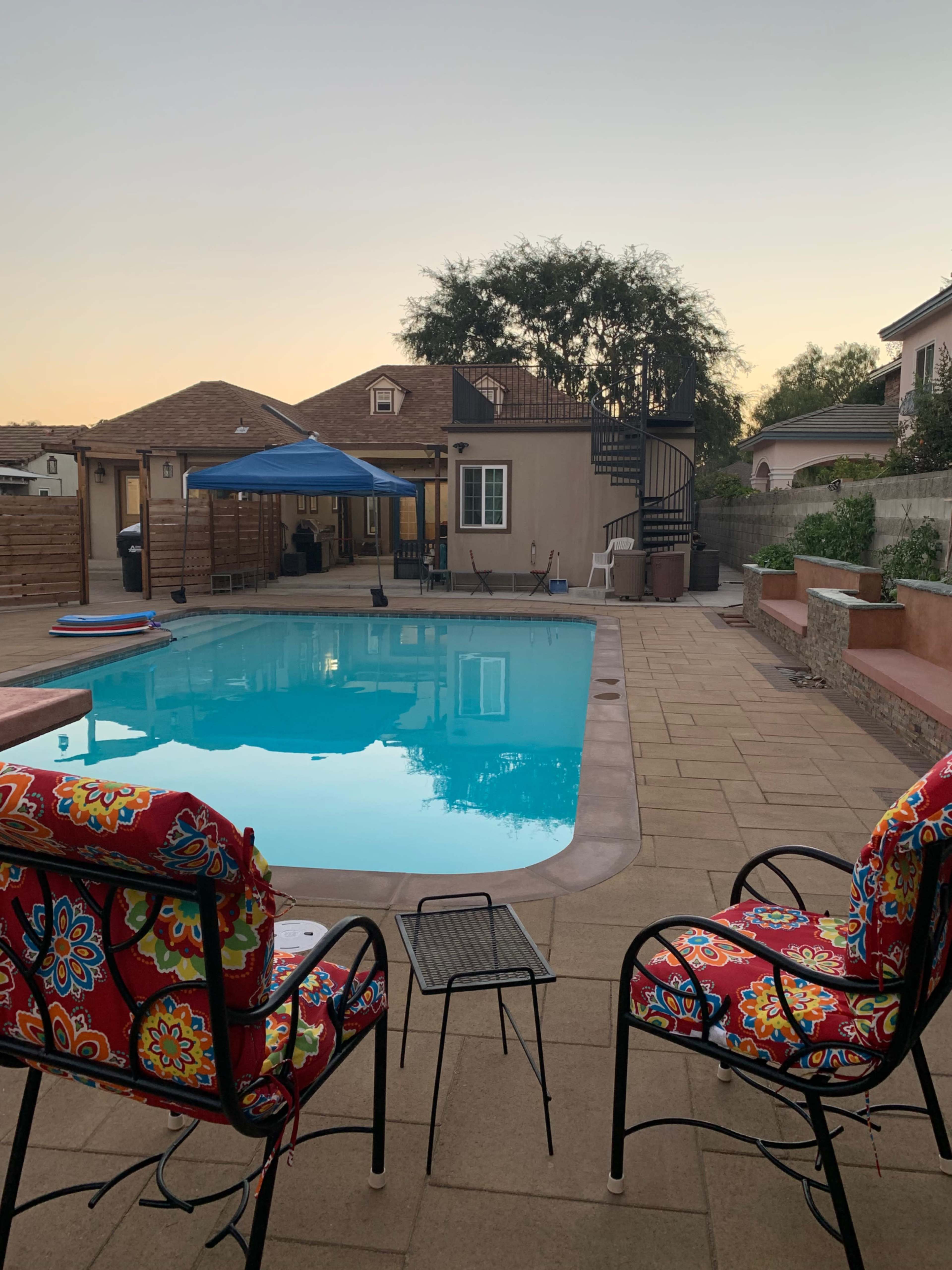 The scene shows a backyard with a swimming pool, two colorful chairs, and a home with a spiral staircase in the background during sunset.