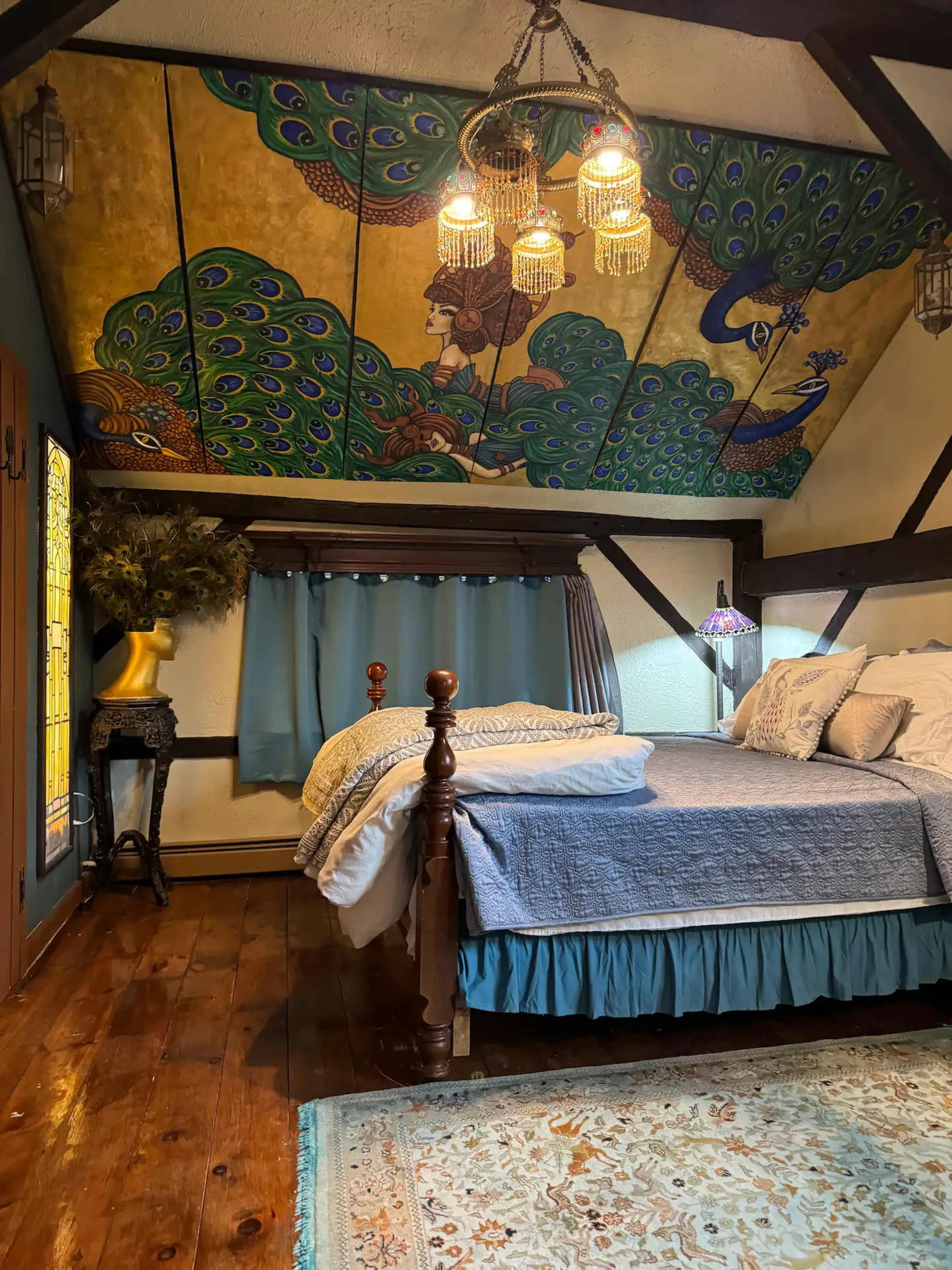 The image shows a cozy bedroom with a wooden bed, a brightly painted ceiling featuring a peacock design, and various decorative elements including a lamp and a vase.