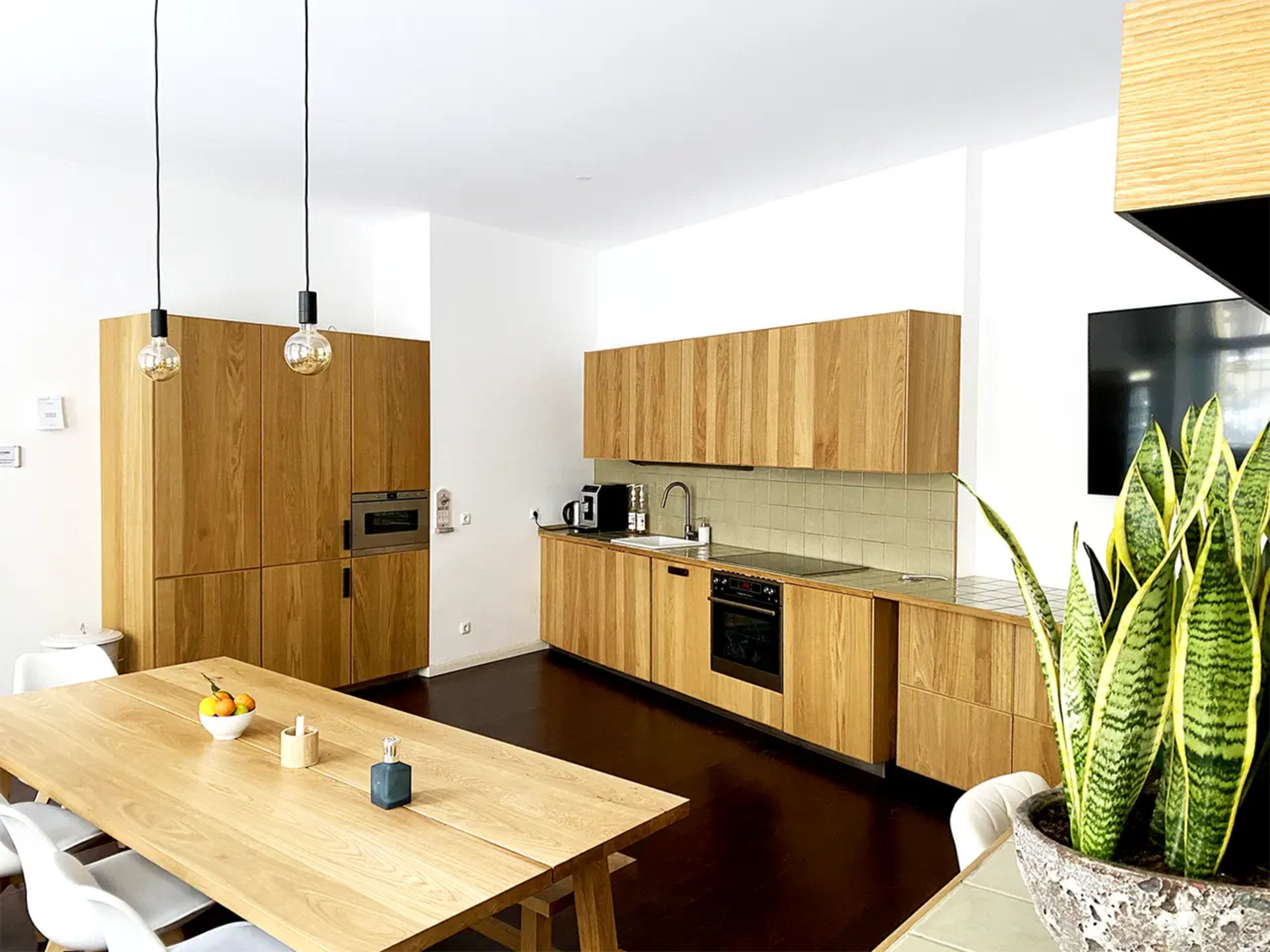The image shows a modern kitchen with wooden cabinetry, a large dining table, and a plant in a corner.