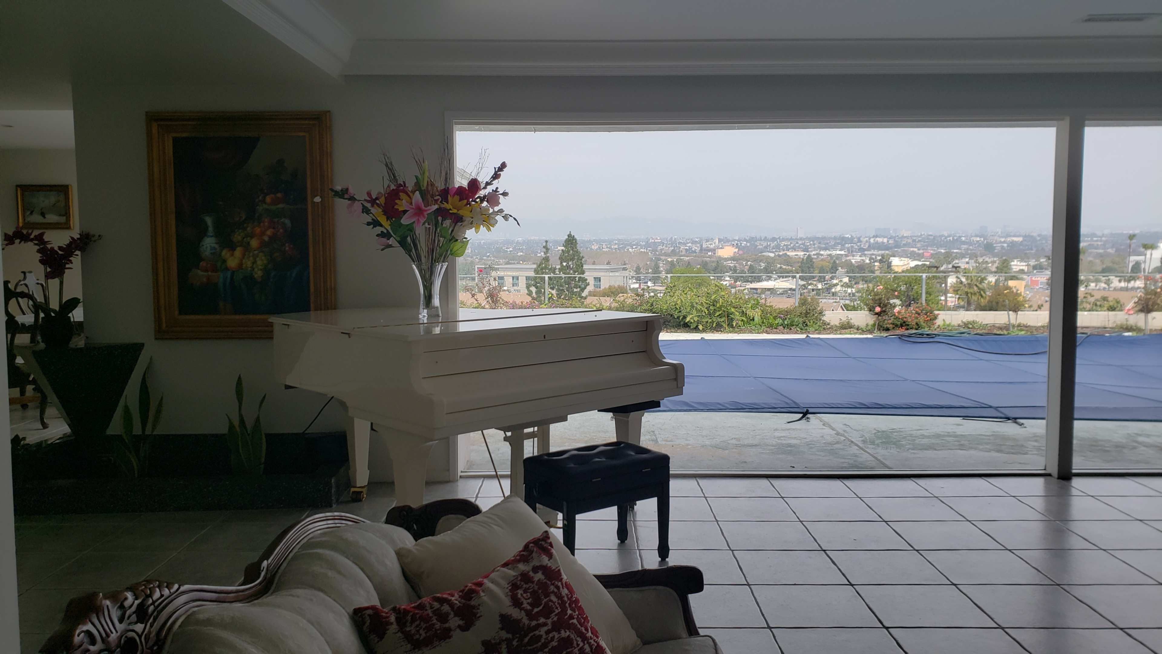 A white piano next to a couch in a room that opens to a view of a city and mountains beyond a covered pool.