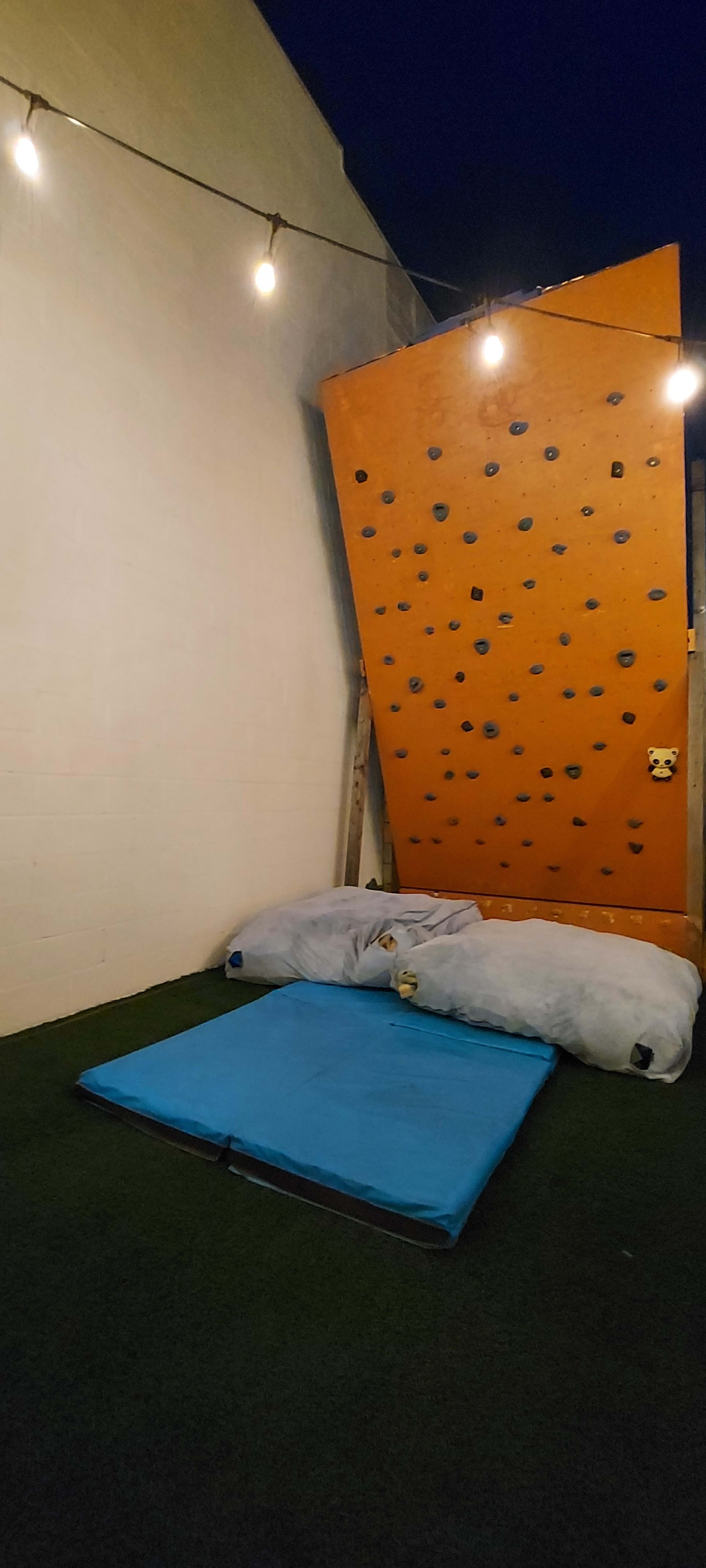 A climbing wall with colorful holds, positioned next to large crash mats on a grassy surface, illuminated by string lights overhead.
